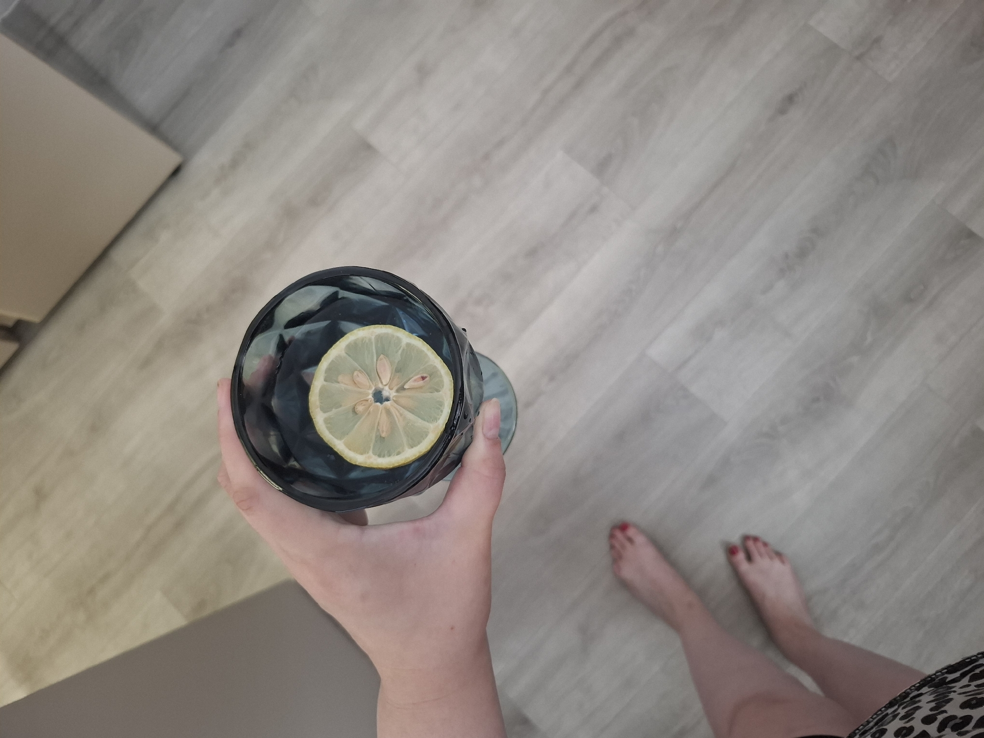 A first-person, overhead perspective shows a hand holding a dark-colored glass with a sliced lemon on top. The person is standing on a light gray wood-patterned floor, and part of their lower legs and feet are visible. The toes are painted red. A cabinet or other piece of furniture is partially visible in the upper left corner of the image. This image can't be verified as being in Амзя, Russia.