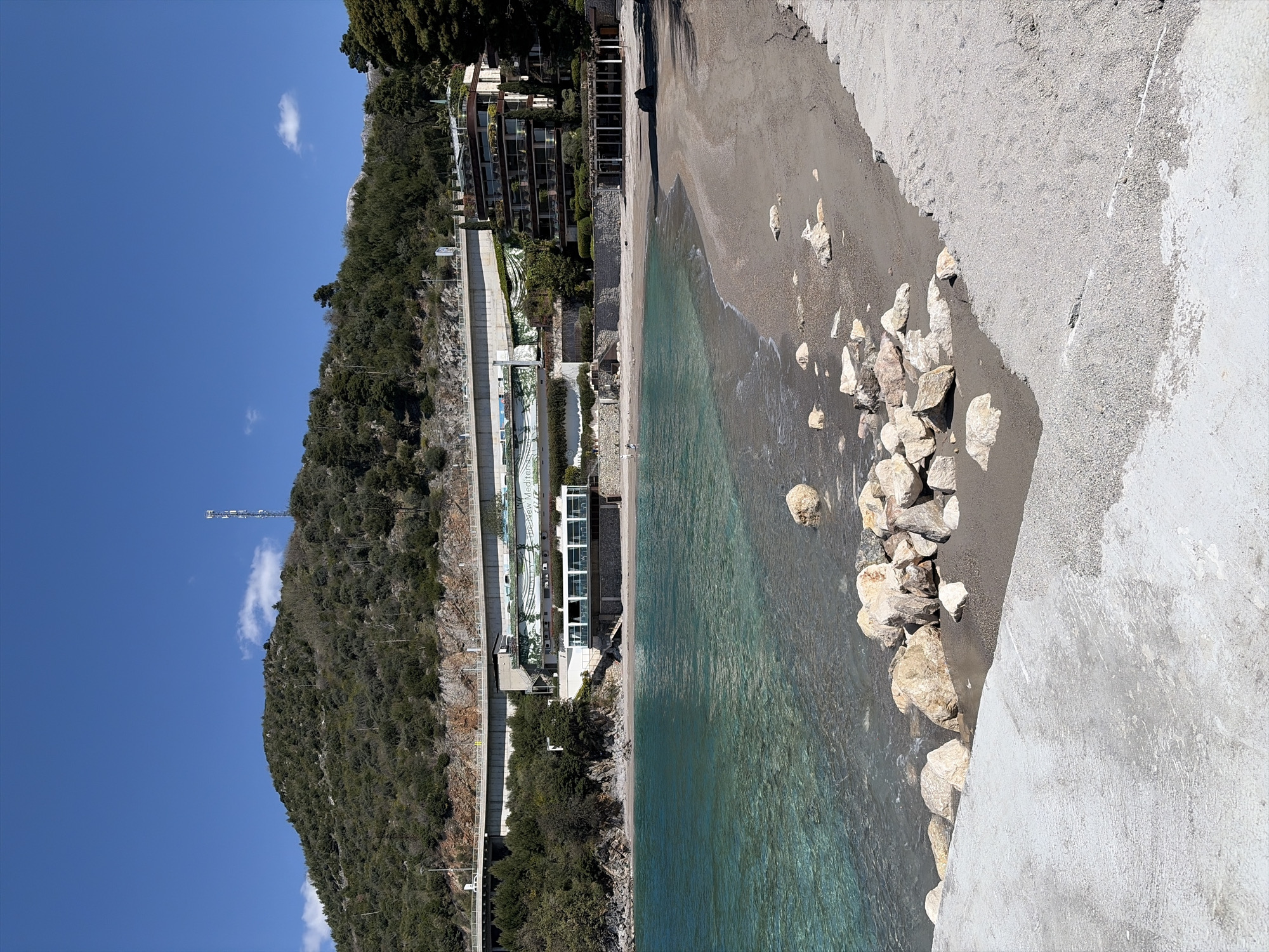 The image shows a beach located in Unknown, Unknown. The beach is characterized by a mix of sand, rocks, and clear water. On the left side of the image, there is a hillside covered with trees, along with buildings and retaining walls along the coast. In the background, there is a communication tower on the hillside. The sky is clear and blue with a few clouds. The foreground shows a concrete structure with rocks piled along the water's edge.