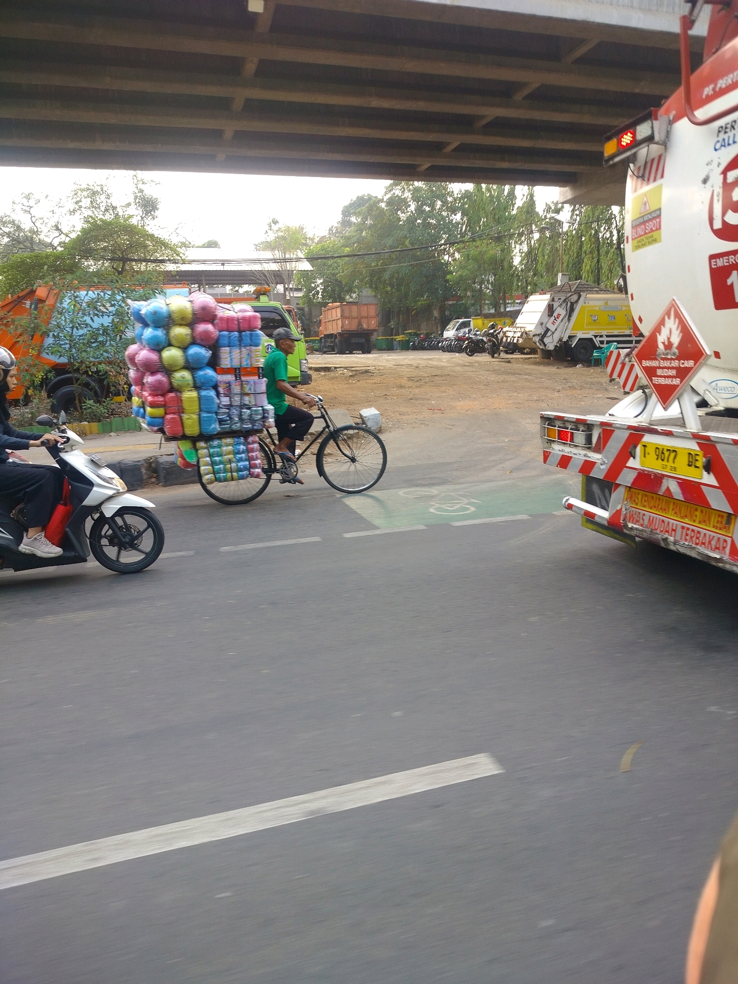 In East Jakarta, Indonesia, a cyclist wearing a green shirt and a cap is riding a bicycle laden with a large cargo of colorful plastic containers, possibly for sale. The cyclist is on a paved road, moving to the right. To the left of the cyclist, a woman on a white scooter, wearing black clothing and a hijab, is also moving to the right. In the background, several vehicles are parked, including a yellow garbage truck and a white tanker truck with hazard markings. The scene is under an elevated road structure, with trees and buildings visible in the distance. The sky is overcast.