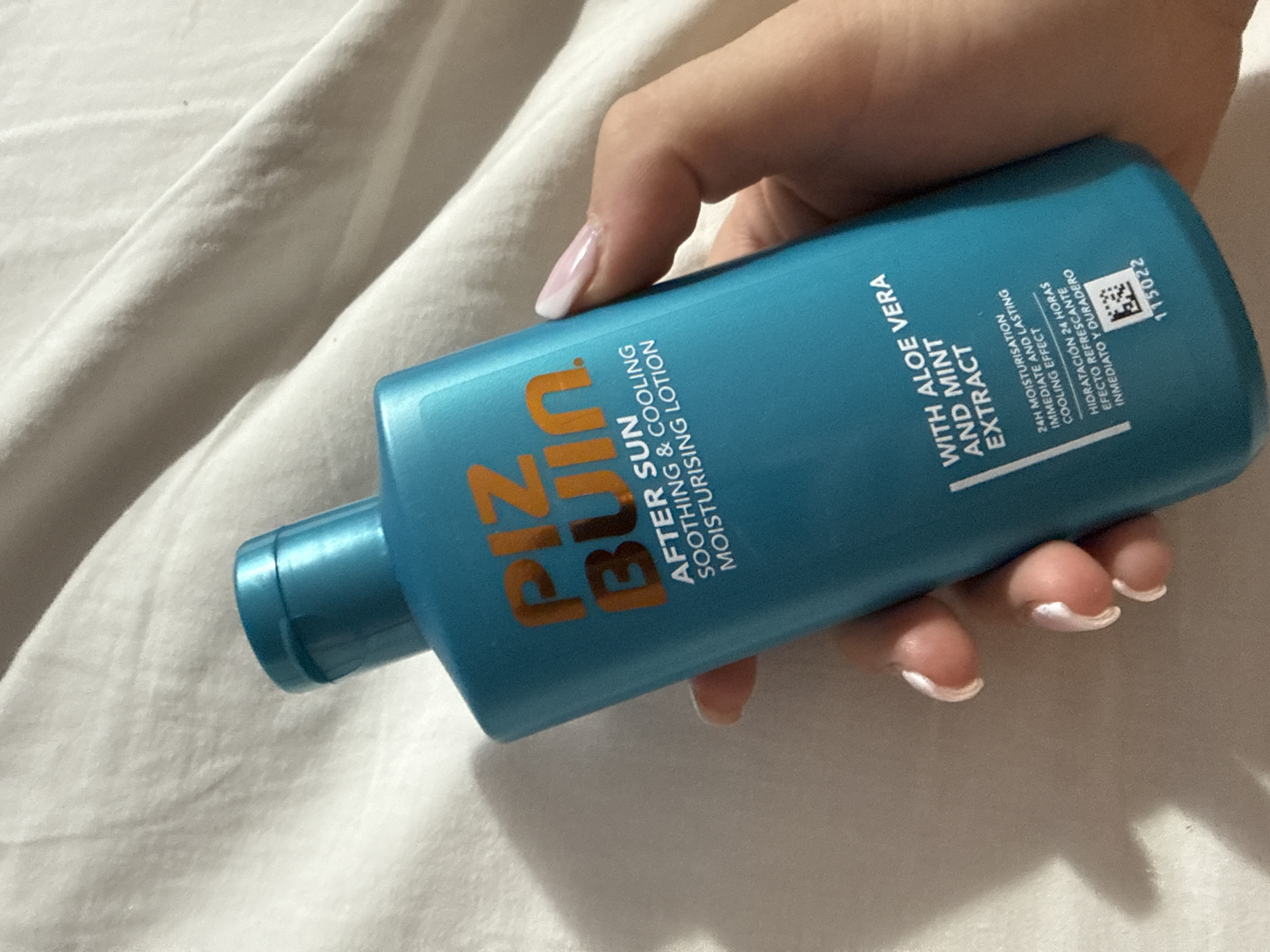 The image shows a hand holding a blue bottle of Piz Buin After Sun Soothing & Cooling Moisturising Lotion. The bottle is labeled "WITH ALOE VERA AND MINT EXTRACT" and includes text in both English and Spanish detailing the product's 24-hour moisturization and cooling effect. The hand has manicured nails with white tips. The background is a white surface, possibly a bed sheet. There is no information in the image suggesting a location in Sofia, Bulgaria.