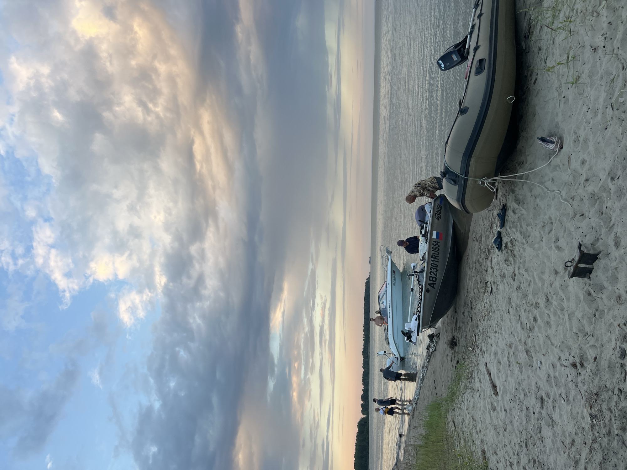 The image depicts a serene scene on a sandy beach in Borovoe, Russia, with several boats moored near the shore. The sky is filled with dramatic clouds, painted in hues of pink, orange, and grey, suggesting either sunrise or sunset. The calm water of the lake stretches towards a distant, tree-lined horizon.
In the foreground, a large, olive-drab inflatable boat lies on the sand, its motor attached. Two pairs of blue sandals are scattered nearby. A smaller, grey aluminum boat is partially submerged in the water, with a white outboard motor mounted on its stern. The license plate of this boat reads "AB2301RUS54" and displays a Russian flag.
Further out in the water, a sleek white motorboat is anchored. It has "Empyrean 590 Lautia" written on its side. Several individuals are visible around these boats. On the shore, near the white boat, four people are standing in the shallow water. One person, wearing a yellow and white striped shirt and dark shorts, is facing away from the camera. Next to them, two individuals in dark shorts are standing. Another person, in a grey t-shirt and dark shorts, is bending over.
In the white boat, two people are visible. One is standing and looking towards the shore. The other, a man wearing a light-colored patterned shirt, is standing near the stern of the aluminum boat, facing the water.
The beach itself is composed of light-colored sand, with some dry seaweed and debris scattered across it. Green grass and reeds grow along the edge of the water. The overall impression is one of a peaceful, end-of-day gathering by the water.
