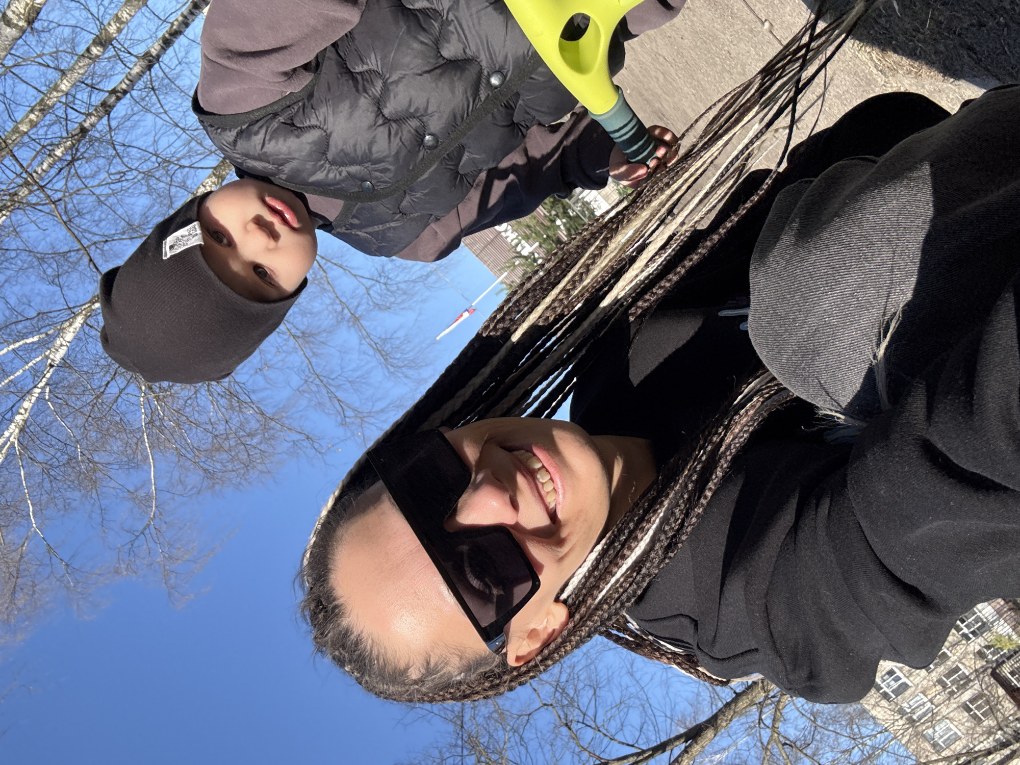 A rotated eye-level medium shot depicts a smiling woman in sunglasses and braided hair facing the camera, along with a young child wearing a black beanie and vest. The woman is dressed in black clothing, with a gray hooded sweatshirt visible. The child holds a neon green scooter or similar wheeled toy. Bare trees are visible, set against a clear blue sky, suggesting a cold weather season. A building with multiple windows and a flag pole is visible in the background. The location is the city of Unknown, Unknown.