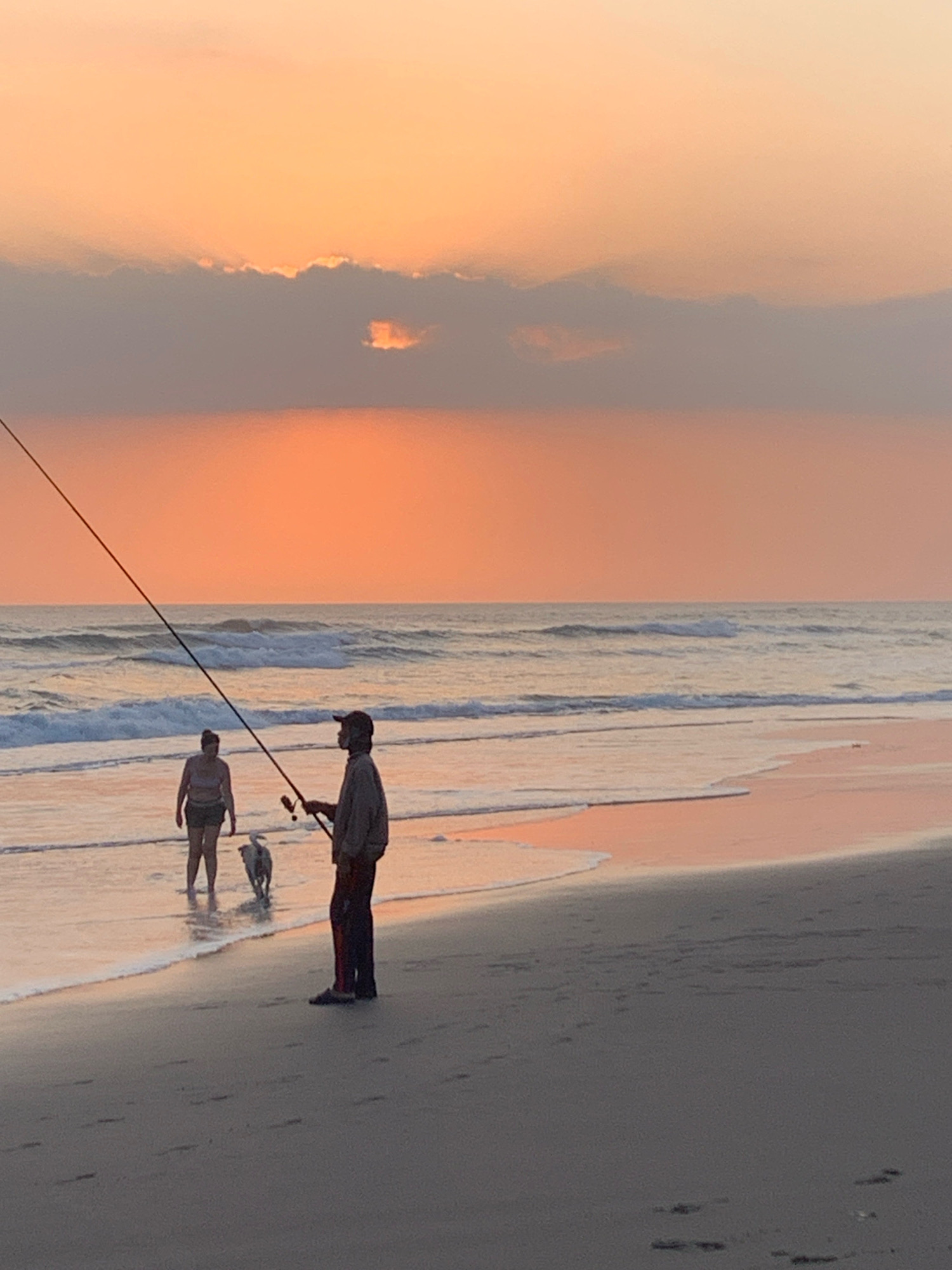 An overcast sunset creates a soft orange glow over a beach scene. In the foreground, a person with a fishing rod stands facing the ocean, while a smaller figure, possibly a dog, plays near their feet. Further out, a woman stands in the shallow water, with a dog that appears to be swimming or playing in the waves. The ocean is calm, with gentle waves lapping at the shore. The sand is wet and reflective in the fading light. There is no indication of the city of Kediri, Indonesia in this image.