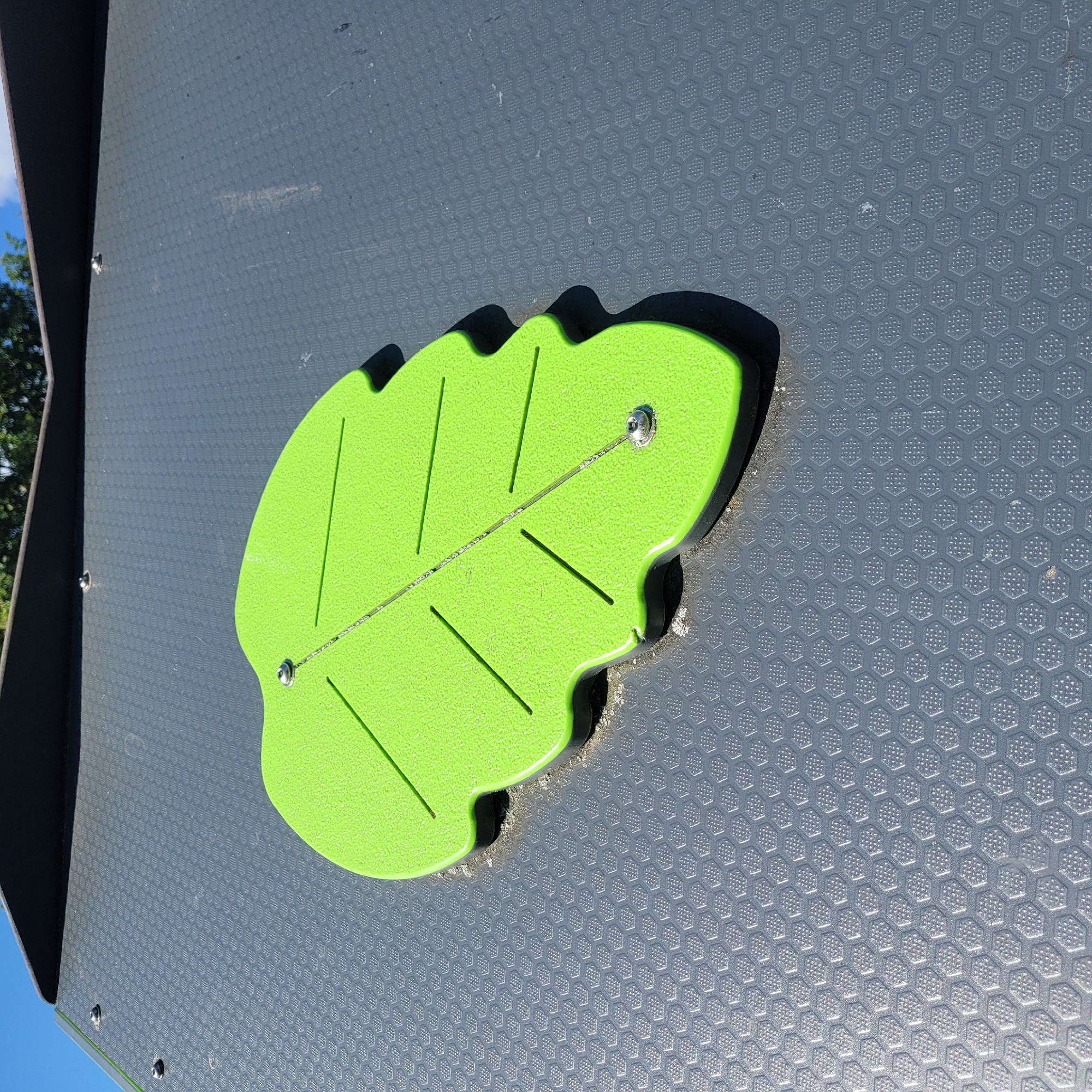 The image shows a close-up, low-angle outdoor shot of a bright green, leaf-shaped climbing hold attached to a dark gray, textured climbing wall. The leaf has three carved veins and is secured to the wall with two visible silver screws. The climbing wall has a hexagonal honeycomb pattern and is part of a larger structure that includes a dark gray overhang at the top of the frame. The background shows a bright blue sky with some wispy clouds and blurred green foliage, suggesting the presence of trees. The overall impression is of a detail on a piece of playground equipment or an outdoor climbing facility, likely located in Jūrmala, Latvia, given the context of the prompt.