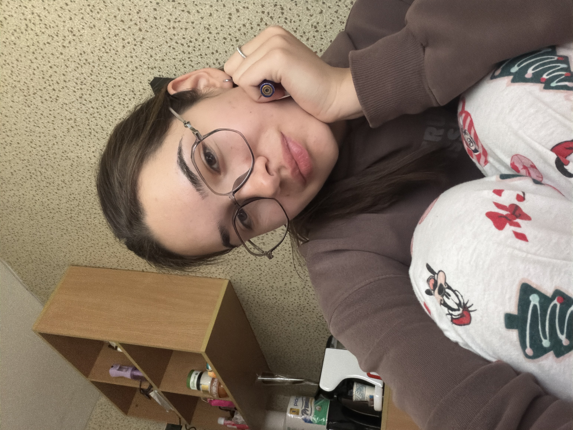 An eye-level indoor portrait captures a young person, likely female, with dark hair and round, thin-rimmed glasses, in a relaxed, reclining pose. Her left hand, wearing a ring, rests near her temple, holding a blue pen. She is dressed in a plain brown long-sleeved top and white pants patterned with festive designs, including green Christmas trees with pink and white ornaments, and a cartoon character resembling Goofy. Her lips are slightly parted, conveying a neutral to pensive expression.
The background features a light brown, textured wall or ceiling. To the left, a light wooden cubby shelf unit is visible, holding various bottles and jars, one with a green label and another partially readable as "PEANUT CREAM." A small purple electronic device is also on the shelf. Below her right arm, a white and red appliance, possibly a mini-fridge or dispenser, is partially visible. The scene suggests a cozy, personal indoor environment, possibly a bedroom, during the holiday season. The location context for the image is Sofia, Bulgaria.