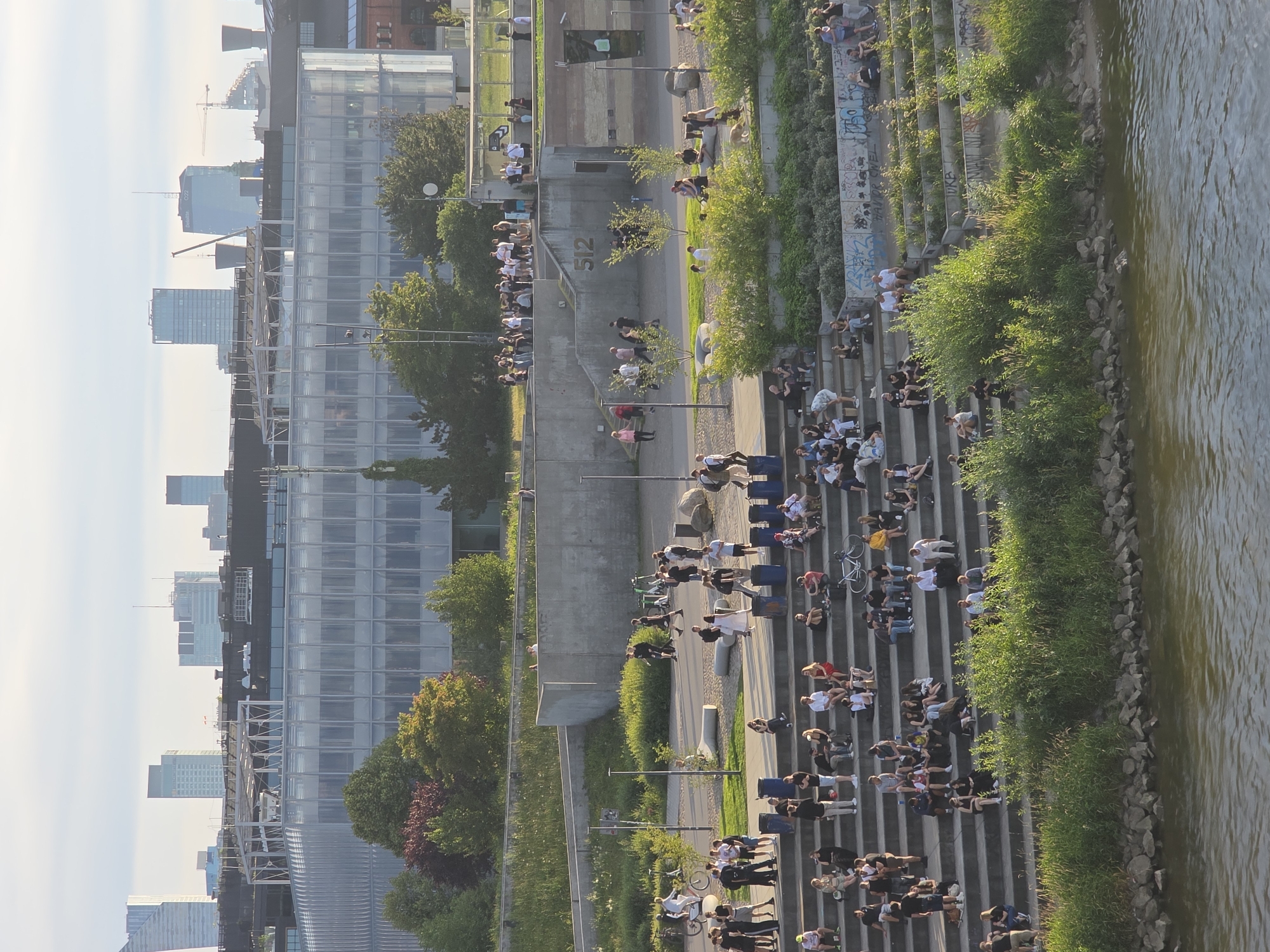 The image depicts a sunny day with a view of the Vistula River embankment in Warsaw, Poland. In the foreground, a body of water flows, with a rocky bank on the right. Behind the bank, a series of stepped terraces rise, populated with numerous people sitting and socializing. These terraces are bordered by lush green vegetation and some low-lying bushes.
Further back, a concrete walkway is visible, with more people walking and cycling. Several blue trash bins are placed along the walkway. Beyond this, a modern glass-fronted building with a distinctive architectural design stands, surrounded by trees. In the background, the skyline of Warsaw is dotted with tall modern skyscrapers. The scene suggests a lively public space, likely a popular spot for leisure and recreation. The sky is mostly clear with a few wispy clouds. The overall atmosphere is that of a pleasant urban environment during daylight hours.