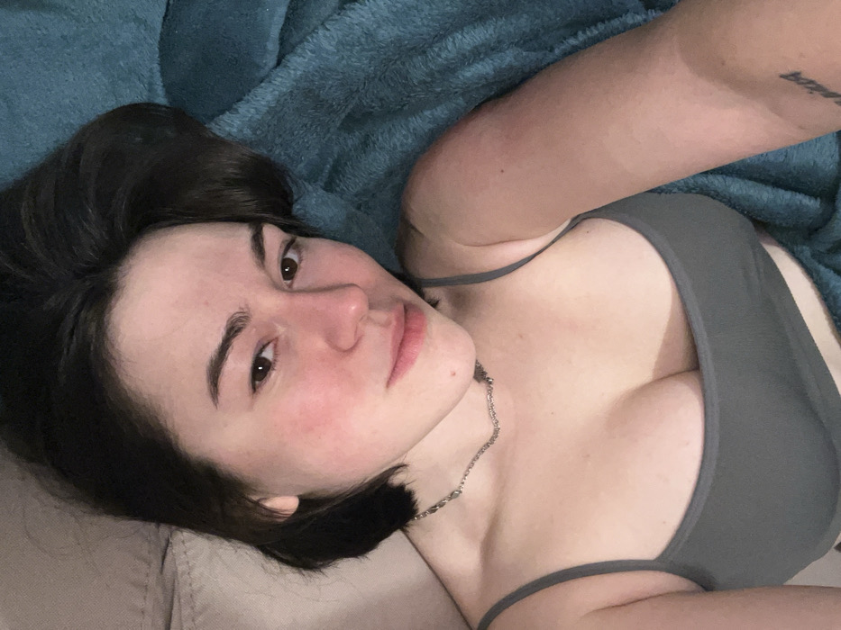 A woman with dark hair and fair skin looks directly at the camera. She is lying down with a blue blanket behind her and a neutral-colored couch beneath her head. She wears a gray tank top and a silver necklace. Her expression is neutral, with a slight smile. The image is a close-up, focusing on her face and upper body, captured from a high angle. A tattoo is visible on her right arm.