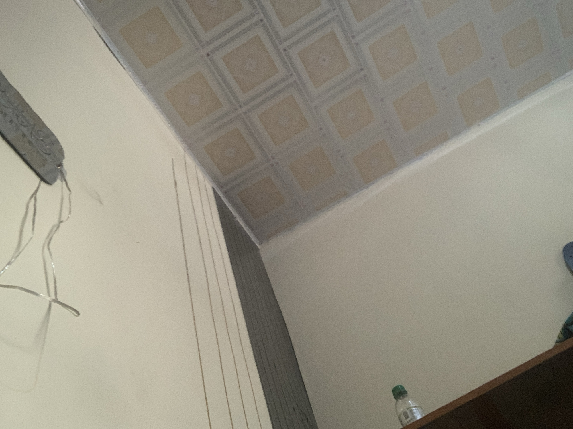 The image depicts an interior scene, likely a room, looking upwards towards a ceiling and walls. The ceiling is paneled with a repeating pattern of light-colored squares, possibly made of vinyl or a similar material, with a grid of darker lines separating them. A metallic object with intricate embossed patterns is visible on the left wall, from which several thin, silver-colored wires hang down. The walls are painted in a pale, neutral color. To the right, a dark wooden shelf is partially visible, on which a clear plastic bottle with a green cap sits. The overall impression is of a simple, possibly residential, interior space in Jauro Yinu, Nigeria.