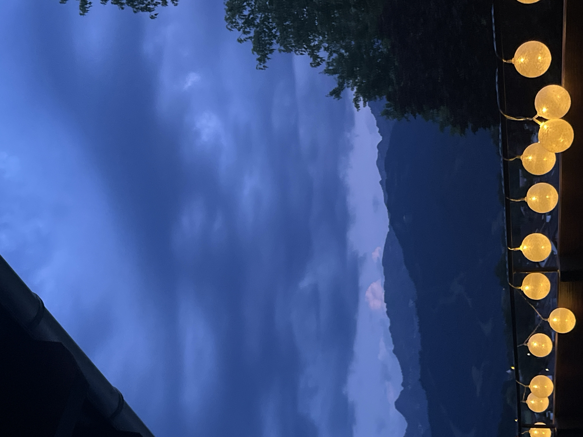 The image depicts a balcony view at dusk in Garmisch-Partenkirchen, Germany. The sky is a deep, cloudy blue, with darker storm clouds gathering. Silhouetted mountain ranges are visible in the distance, with faint hints of lighter cloud formations over the peaks. In the foreground, a string of spherical, illuminated lights hangs from a railing. These lights are a warm, yellow hue and appear to be made of a translucent material. To the right, the dark silhouette of a large tree with dense foliage is visible against the twilight sky. The overall impression is one of impending weather and a serene, yet slightly dramatic, evening landscape.