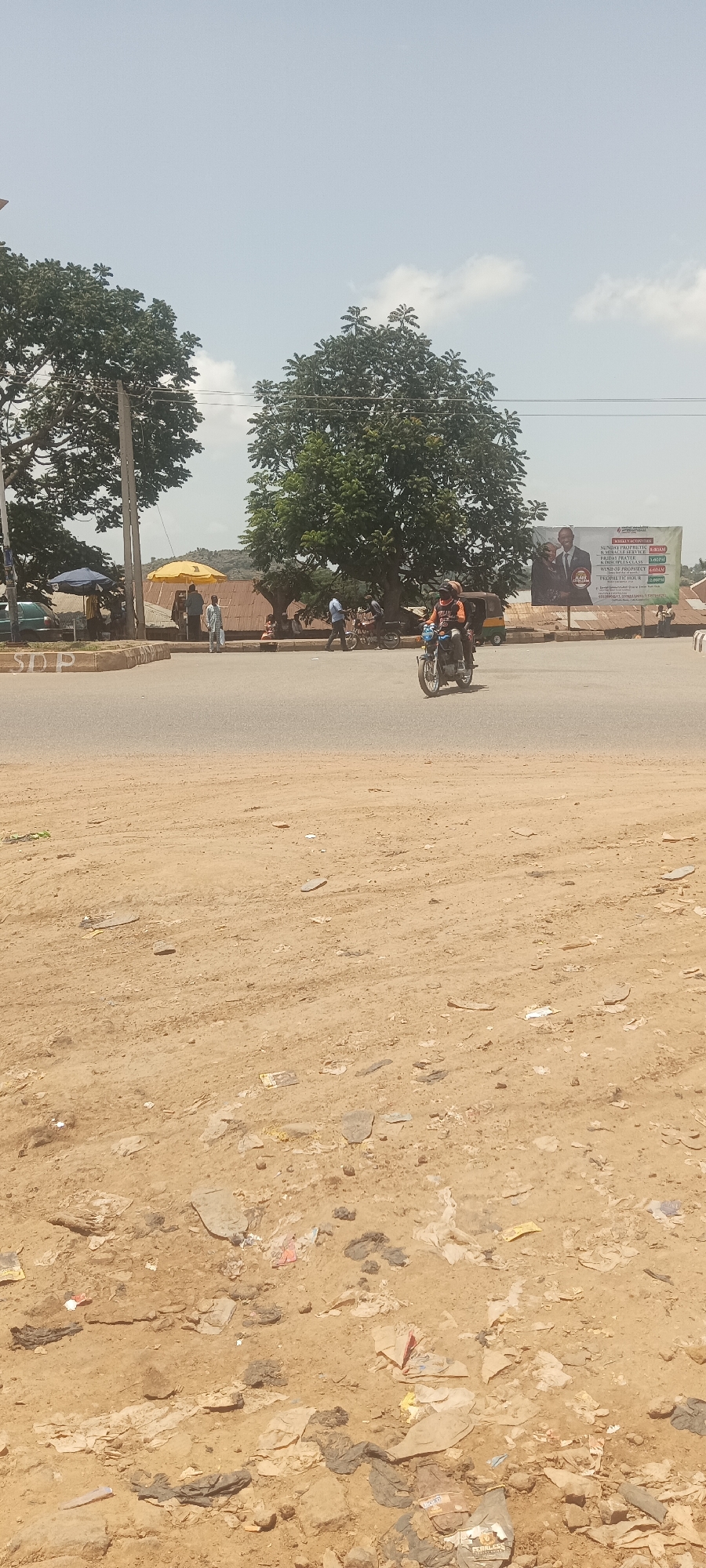 A street scene in Bukuru, Nigeria, shows a motorcycle carrying two people moving across a paved road. The foreground is a dirt area with scattered litter. In the background, there are trees, buildings, and people gathered near a yellow umbrella. A billboard with portraits and text is visible to the right.