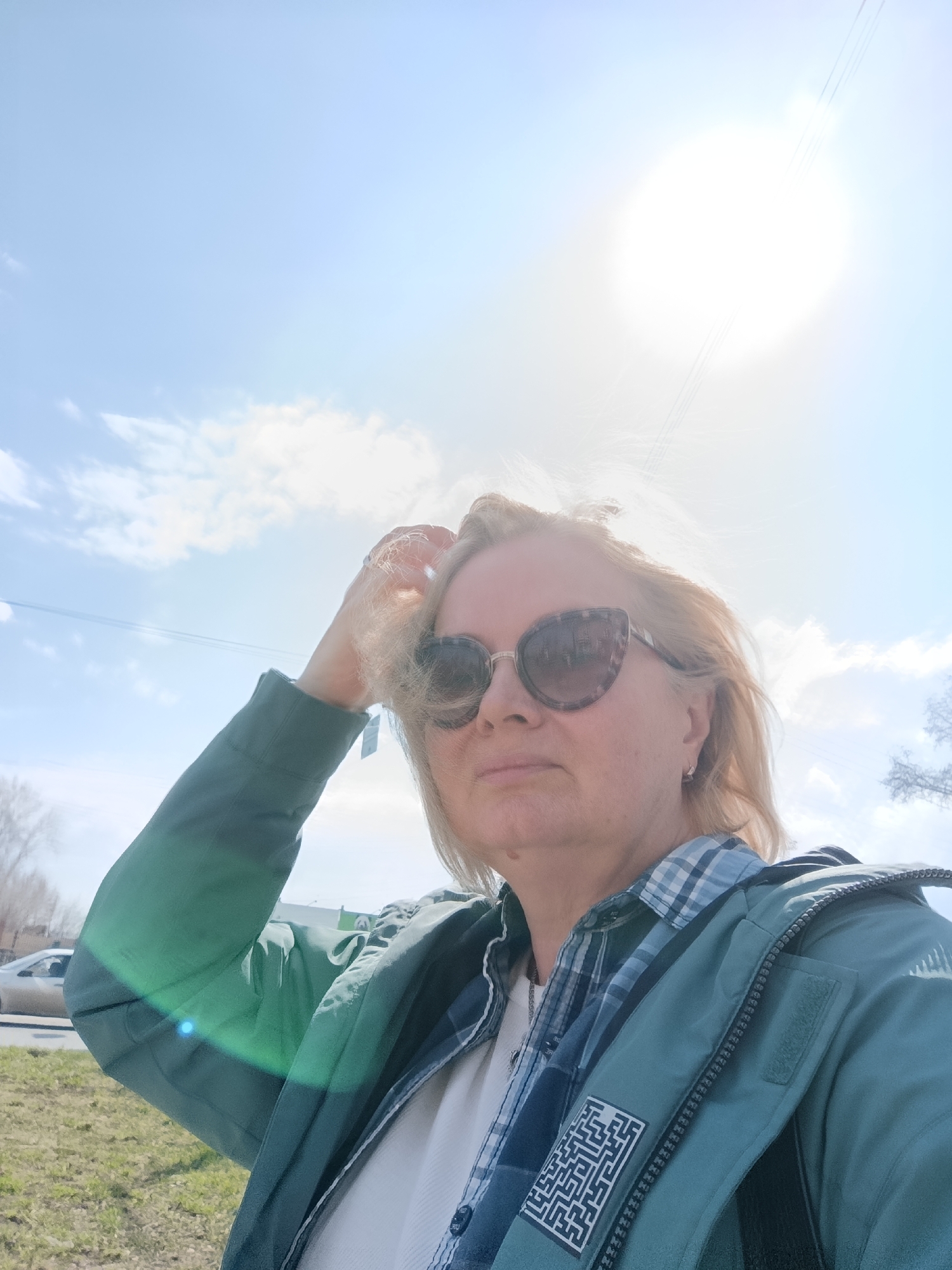 A fair-skinned woman with blonde hair, wearing sunglasses and a teal or greenish-blue jacket over a blue and white plaid shirt, takes a selfie outdoors in Chaikovsky, Russia. She holds her right hand to her hair, which appears slightly windblown. Her expression is neutral, partially obscured by large, cat-eye style sunglasses with a brown and clear pattern. Small, silver-colored hoop earrings are visible on her ears.
The scene is set on a very bright, sunny day, indicated by the prominent sun in the upper right, causing significant lens flare and overexposure in that area of the sky. The sky is clear blue with some scattered white clouds. A road with a light-colored car and a grassy verge are visible in the lower left background, along with bare trees, suggesting late winter or early spring. Utility lines or power lines stretch across the sky in the mid-ground.
On the left sleeve of her jacket, a black and white rectangular patch displays a complex maze pattern. No readable text is visible.