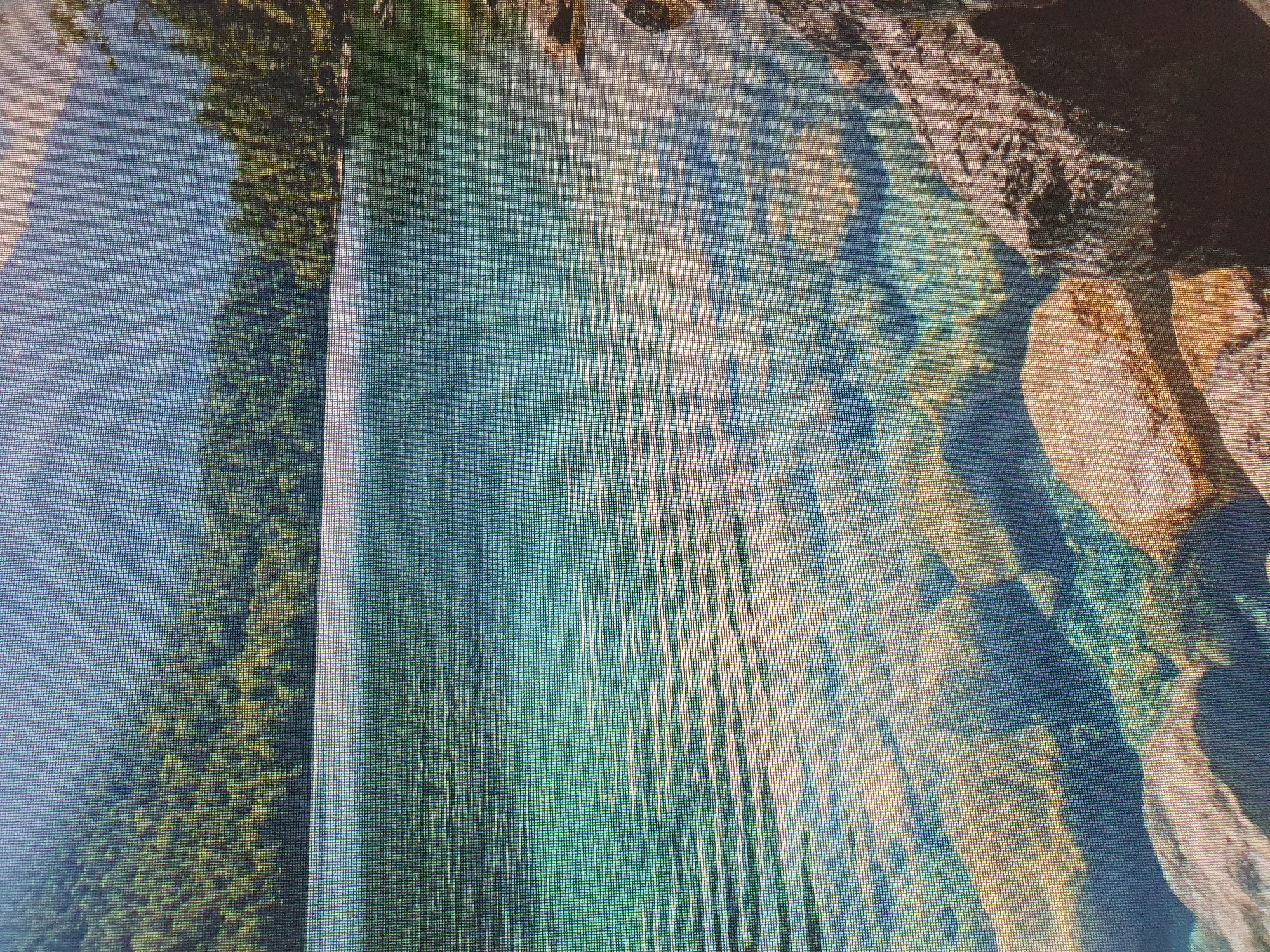 This is a photograph of a clear, turquoise lake with visible rocks and pebbles on its bed. In the background, a dense forest of green trees covers a sloping mountainside, leading up to a hazy, blue sky. The water has gentle ripples, suggesting a calm atmosphere. The foreground shows large, light-colored rocks partially submerged in the water along the shoreline. The overall scene depicts a serene natural landscape. There are no people or man-made structures visible in the image. The photograph is taken from a low angle, looking across the water towards the forest and mountains. There is no information in the image to suggest this location is in Tashkent, Uzbekistan.