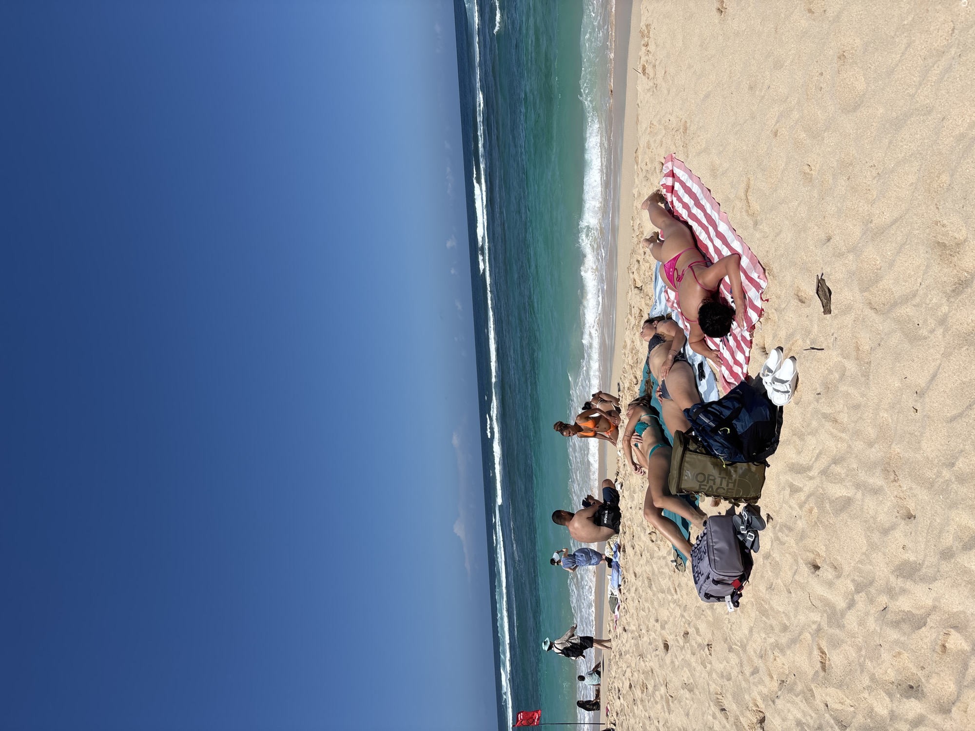 A group of people are sunbathing on a sandy beach in Kuta Selatan, Indonesia, with the turquoise ocean and white-capped waves in the background under a clear blue sky. In the foreground, four individuals are lying on towels on the sand. One person, wearing a teal bikini, is on their back with their eyes closed. Next to them, another person, also on their back, is partially visible. Further down, a person in a pink bikini is lying on their stomach. Between them are several bags, including a gray backpack and a green backpack, along with some sandals and a towel. In the middle ground, more people are visible sitting and standing near the water's edge. A red flag is planted in the sand on the left side of the image. The beach appears to be a popular spot for recreation and relaxation.