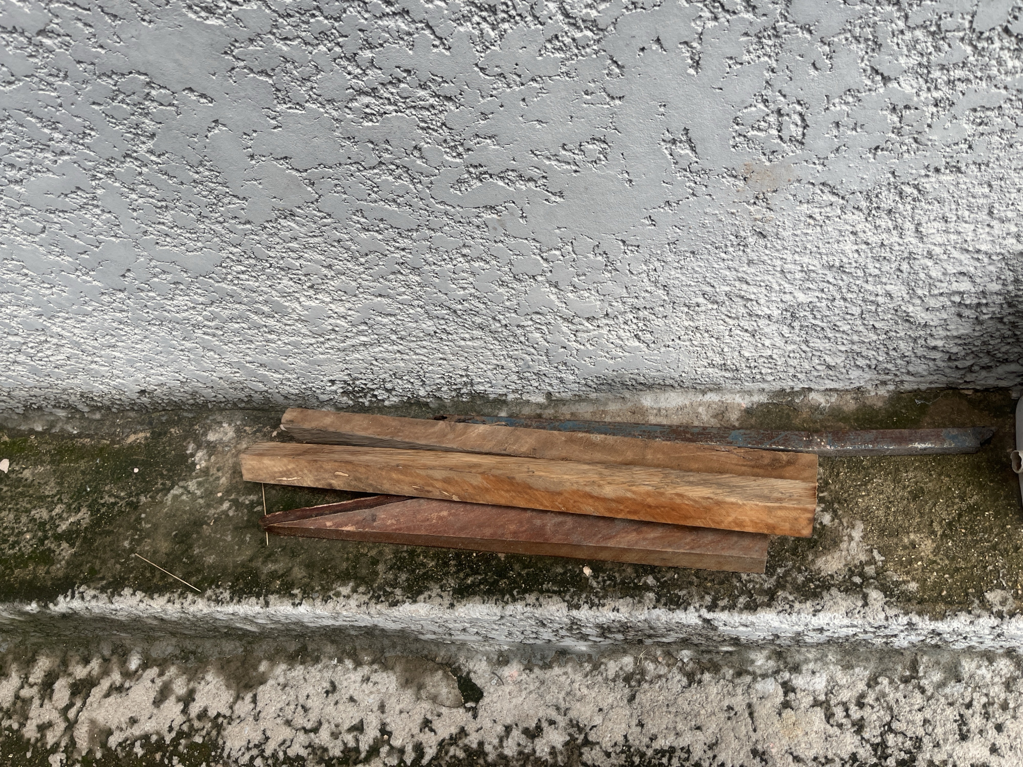 In Jalingo, Nigeria, a close-up, overhead shot captures a textured, light gray concrete wall above a weathered outdoor concrete step. The wall's surface is rough and uneven, showing areas of shadow and light. Below, the concrete step exhibits a gritty, speckled texture with patches of green moss and some white efflorescence. A small pile of four wooden planks is arranged on the step. Two of the planks are a lighter, reddish-brown wood, while the upper two appear to be of a similar wood type but with a darker, richer brown hue and visible grain. One plank on the far right has a rusty blue metallic piece attached to its end. A thin, light-colored stick lies diagonally on the step. The scene appears to be outdoors, possibly in an alleyway or a less-maintained outdoor area.