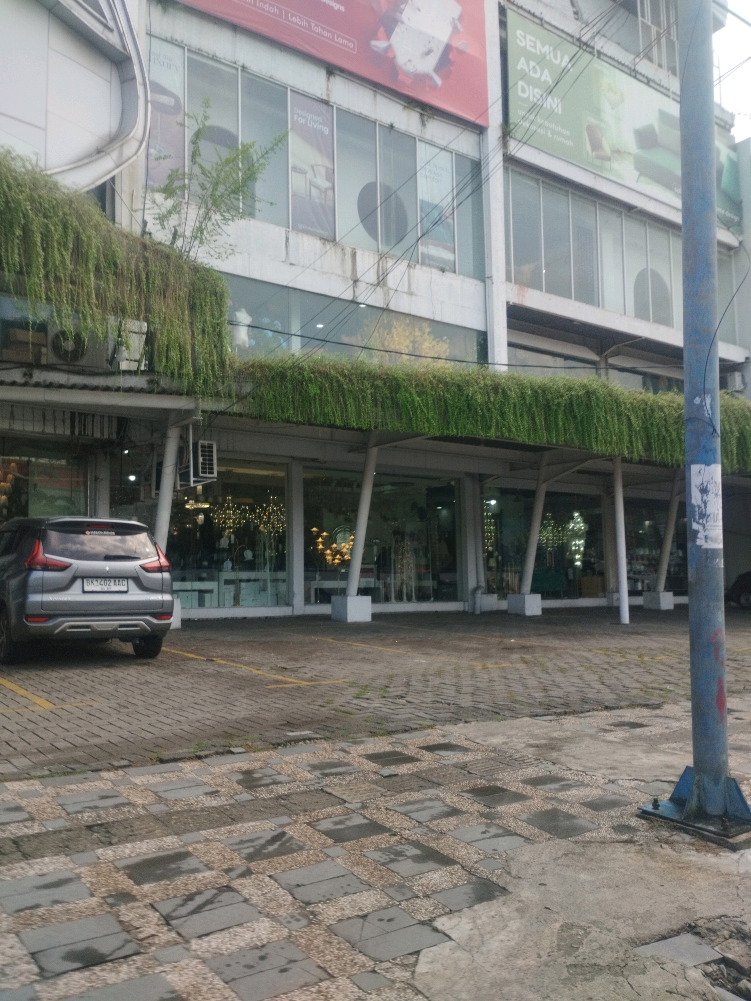 A grey SUV is parked in front of a modern building with large glass windows. The building has a second floor with a green banner that says "SEMUA ADA DI SINI". There are hanging plants on the exterior of the building, creating a green overhang. The ground in front of the building is paved with interlocking stone tiles. A blue metal pole stands to the right of the frame. The scene appears to be an outdoor commercial area in the City of Medan, Indonesia.