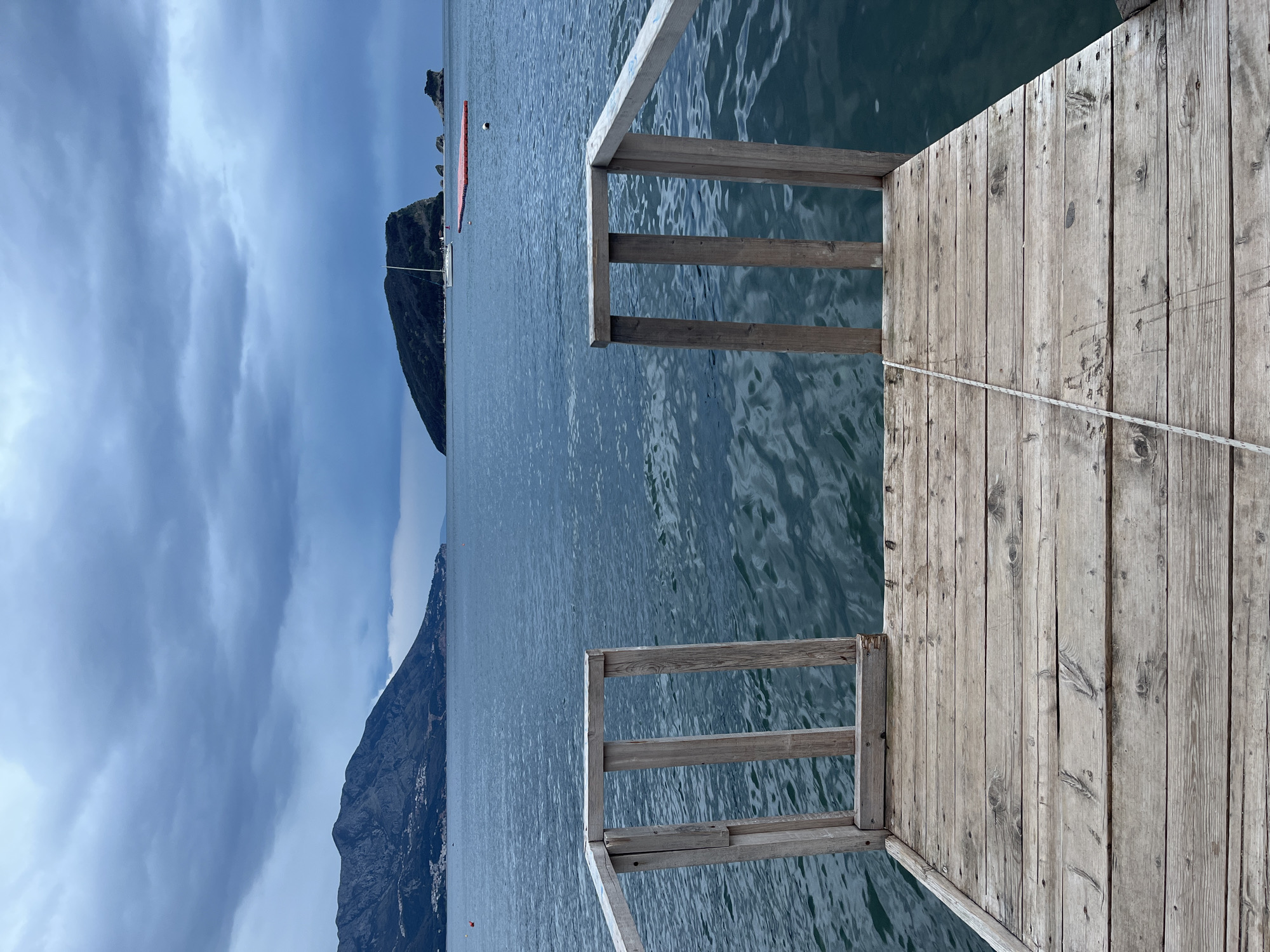 The image shows a pier extending into a body of water, likely the sea. The pier is made of wooden planks with wooden railings. A rope is tied to the pier. In the background, there are two mountainous landmasses visible, partially obscured by cloud cover. There is also a small red inflatable platform visible in the water. The location is likely Budva, Montenegro, considering the typical coastal landscape.