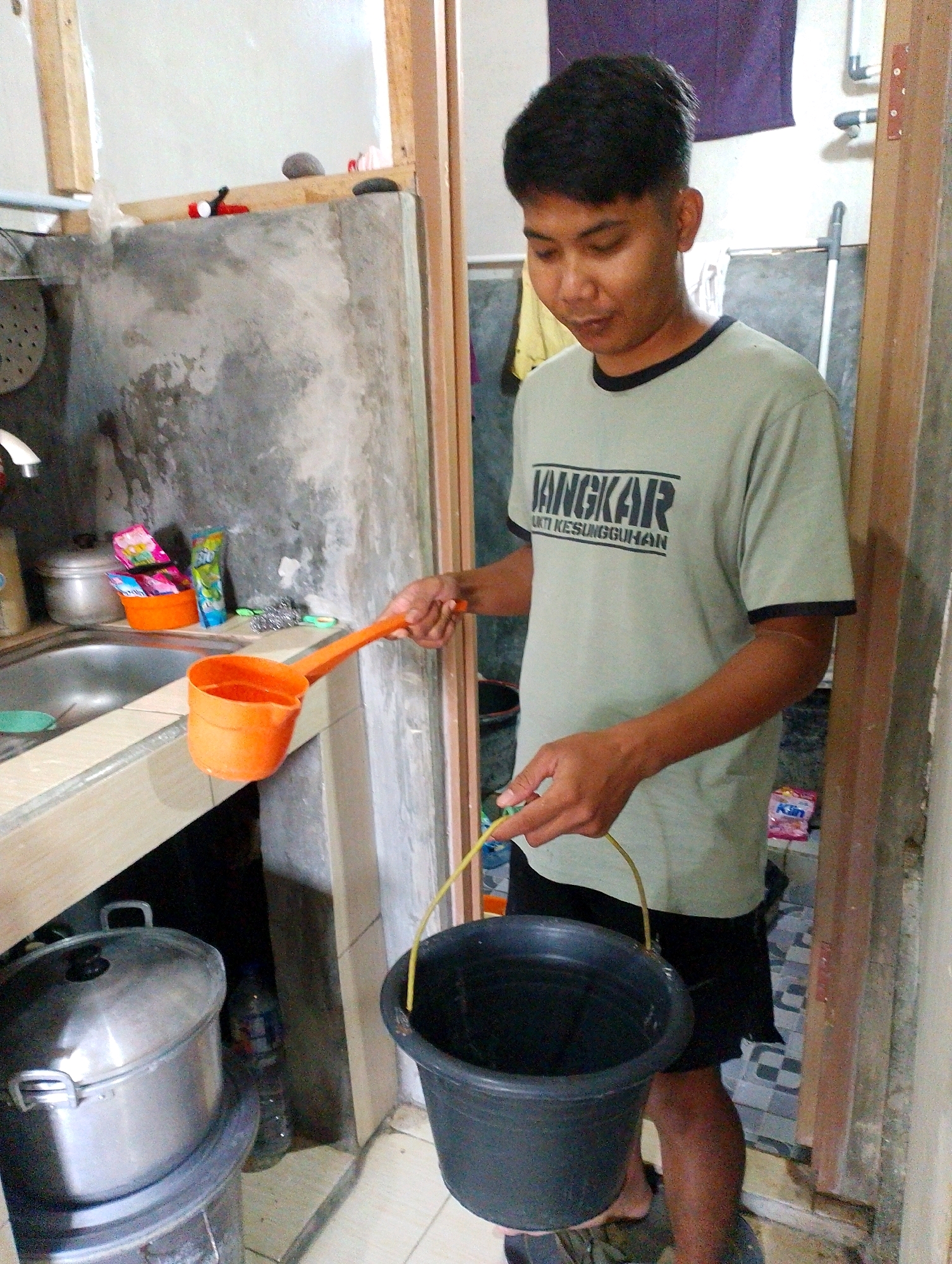 In Desa Babakan, Indonesia, a young adult male stands in what appears to be a utilitarian kitchen or utility area. He is wearing a light green t-shirt with text and black trim, and black shorts. In his right hand, he holds a long-handled orange water dipper. His left hand is shown holding the handle of a black plastic bucket which is partially filled with water. The background includes a concrete wall, a sink with a faucet, various containers, and wooden structures. The overall impression is of a domestic or service setting where water is being manually transported.