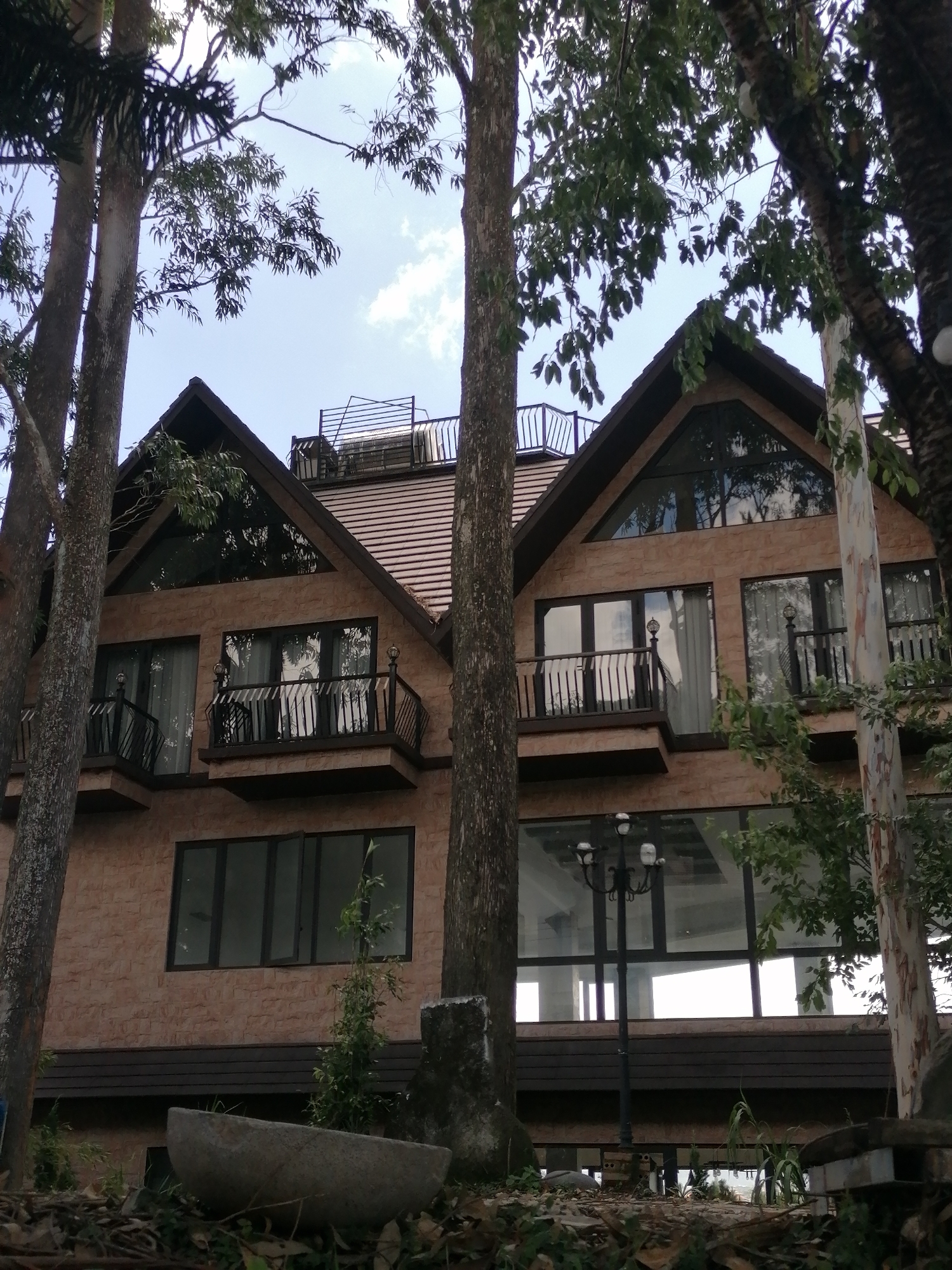 The image shows a multi-story building with a light brown brick facade, a dark brown tiled roof, and large windows. The building features several balconies with black railings and decorative finials. The architecture suggests a modern design with some traditional elements, possibly a hotel or a residential complex. The building is partially obscured by several large trees with lush green foliage, indicating a natural or park-like setting. In the foreground, a large stone trough or planter is visible amidst fallen leaves and vegetation, further suggesting an outdoor environment. A street lamp with multiple globes stands near the building's entrance. The sky above is partly cloudy. While the image doesn't explicitly show the city of Da Lat, Vietnam, the presence of lush vegetation, a large building with potentially resort-like architecture, and a somewhat temperate climate suggested by the foliage are consistent with the characteristics of Da Lat.