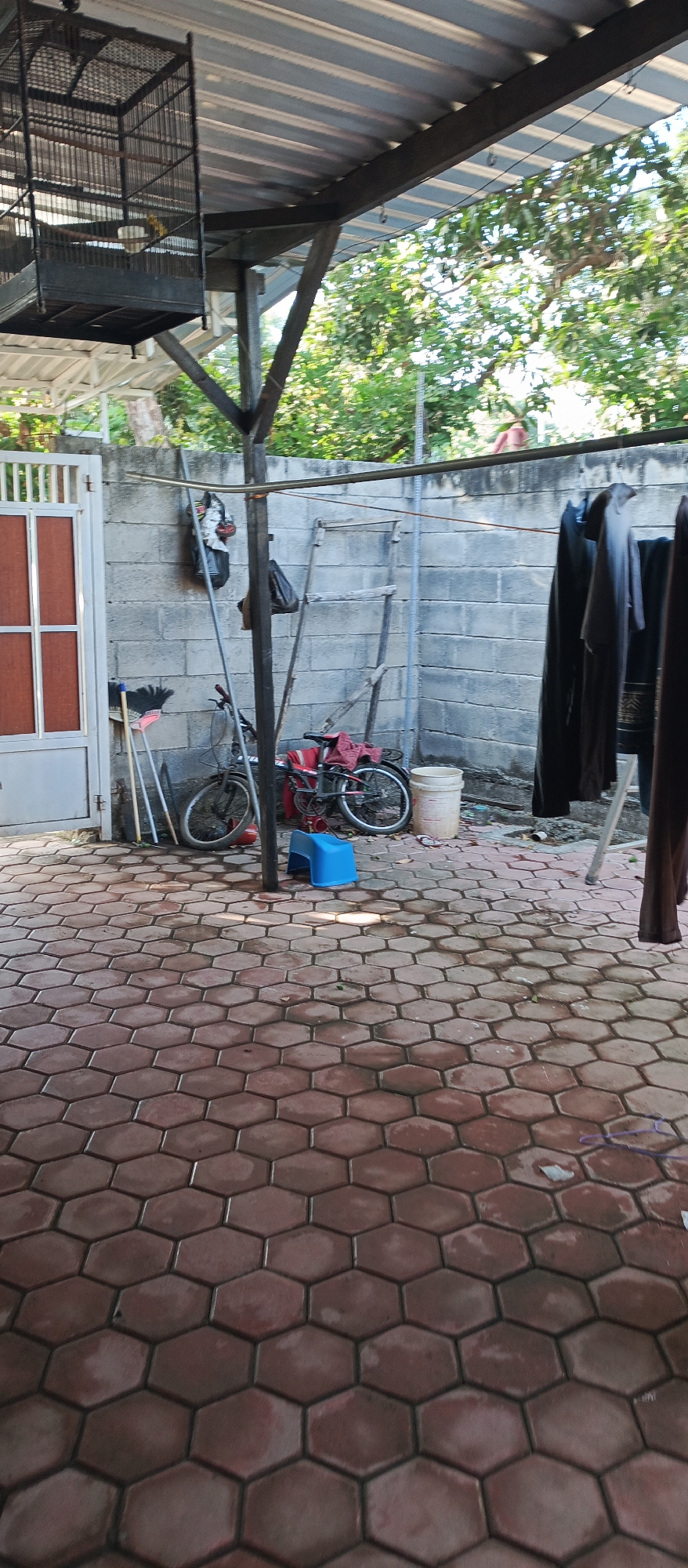 The image depicts a backyard or side yard of a building in Arjasa, Indonesia. The ground is paved with hexagonal red bricks. To the left, a white gate with vertical bars and a reddish-brown rectangular panel is visible. Adjacent to the gate, a broom with a black brush and wooden handles leans against a wall.
Further into the yard, a gray brick wall forms the backdrop. Various items are leaning against or hanging on this wall, including what appears to be bags and a ladder-like structure. Two bicycles are positioned against the wall; one is dark-colored with red accents and a towel on the seat, and another is partially obscured. A small blue plastic stool is placed on the ground near the bicycles.
A clothesline runs horizontally across the scene, with dark-colored garments hanging from it, suggesting laundry is being dried. A large birdcage made of black metal is suspended from the overhang of a corrugated metal roof. The roof is supported by dark-colored metal beams. Beyond the wall, green foliage and trees are visible, indicating an outdoor, possibly tropical, environment. The lighting suggests it is daytime. There are no people clearly visible in the image.