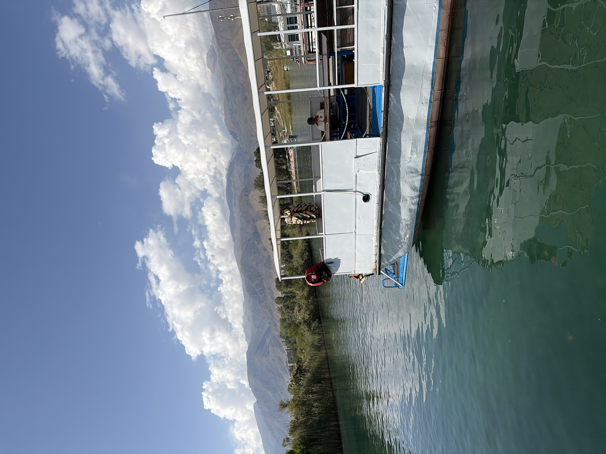 A white passenger boat with a blue trim is docked on a body of water, possibly Issyk-Kul Lake in Cholpon-Ata, Kyrgyzstan. The boat has an open-air seating area with tables and benches, covered by a white canopy. A person wearing a white jacket and a dark cap is seated at one of the tables, looking down. A red life vest is hanging on the railing of the boat. The water is a clear, dark green, reflecting the sky and the boat. In the background, a lush green shoreline with tall reeds and trees is visible, leading to a range of rugged, dry mountains under a partly cloudy blue sky. The overall scene depicts a calm day on the water, with the boat appearing to be ready for passengers or having just completed a tour.