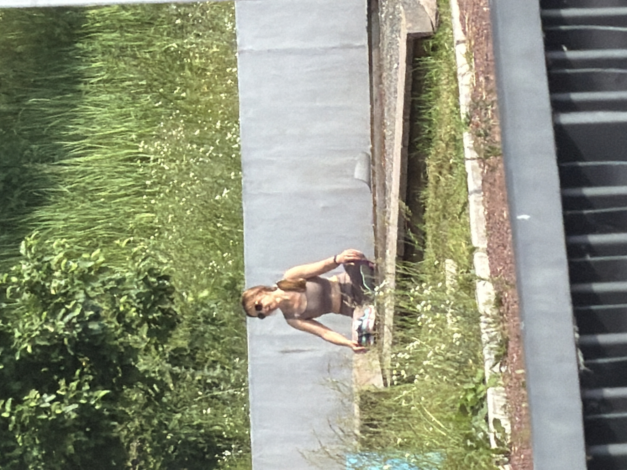 A woman is sitting in a cross-legged pose on a wooden platform. She is wearing a beige tank top and matching pants, with sunglasses on her face and her hair pulled back in a ponytail. To her left, on the platform, is a closed backpack. The platform is situated outdoors, with a gray concrete wall behind it. The area is surrounded by lush green grass and vegetation, with some small white flowers scattered throughout. The scene appears to be in a park or natural setting, potentially in Chaikovsky, Russia, given the context of the request.