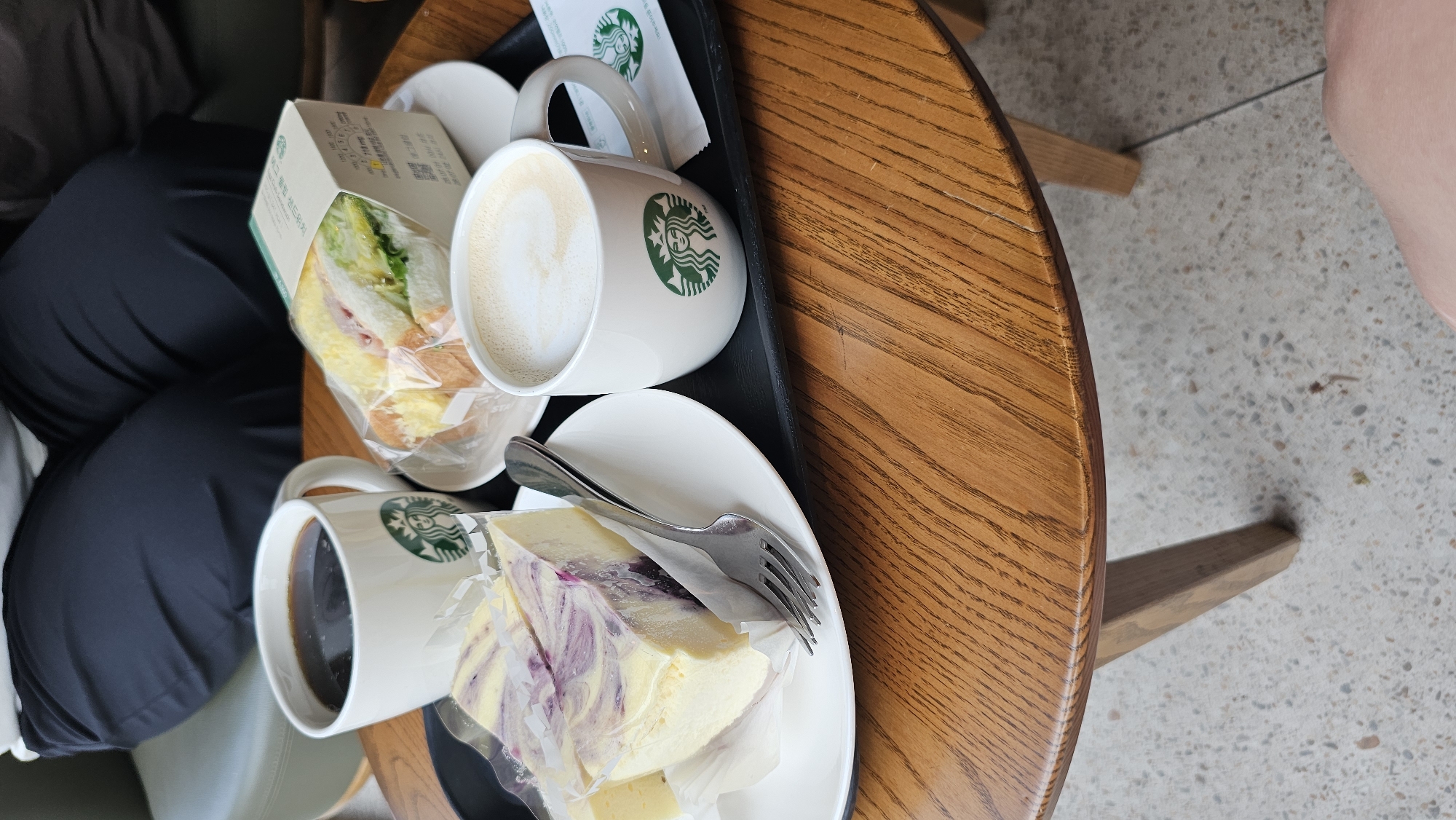A close-up, eye-level shot shows a black tray on a wooden table. On the tray are two Starbucks mugs, a slice of cheesecake, and a sandwich in a box. The mug on the left contains black coffee. The mug on the right contains latte art. The cheesecake is a light yellow with purple swirls and is wrapped in plastic. The sandwich appears to be made with white bread, egg, ham, and lettuce. The box for the sandwich is white and has Korean text. A fork rests on the plate with the cheesecake. In the background, a person wearing dark pants sits on a green chair. The flooring is speckled concrete. This scene is likely at a Starbucks in Suwon-si, South Korea, given the presence of Starbucks branded items and the inferred location.