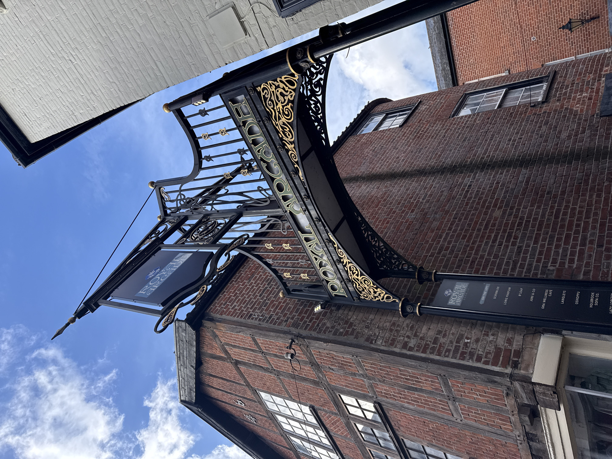The image shows the entrance to the Royal Arcade in Norwich, United Kingdom. The arcade is marked by an ornate black and gold metal structure, with a sign that lists businesses within the arcade, including "Scribbler," "Loris Parfum," "B'Leaf," and "Lazy Lobster." The building is brick with some timber framing visible on the lower portion. The sky is visible between the buildings, with some clouds. The photo is taken from a low angle looking up.