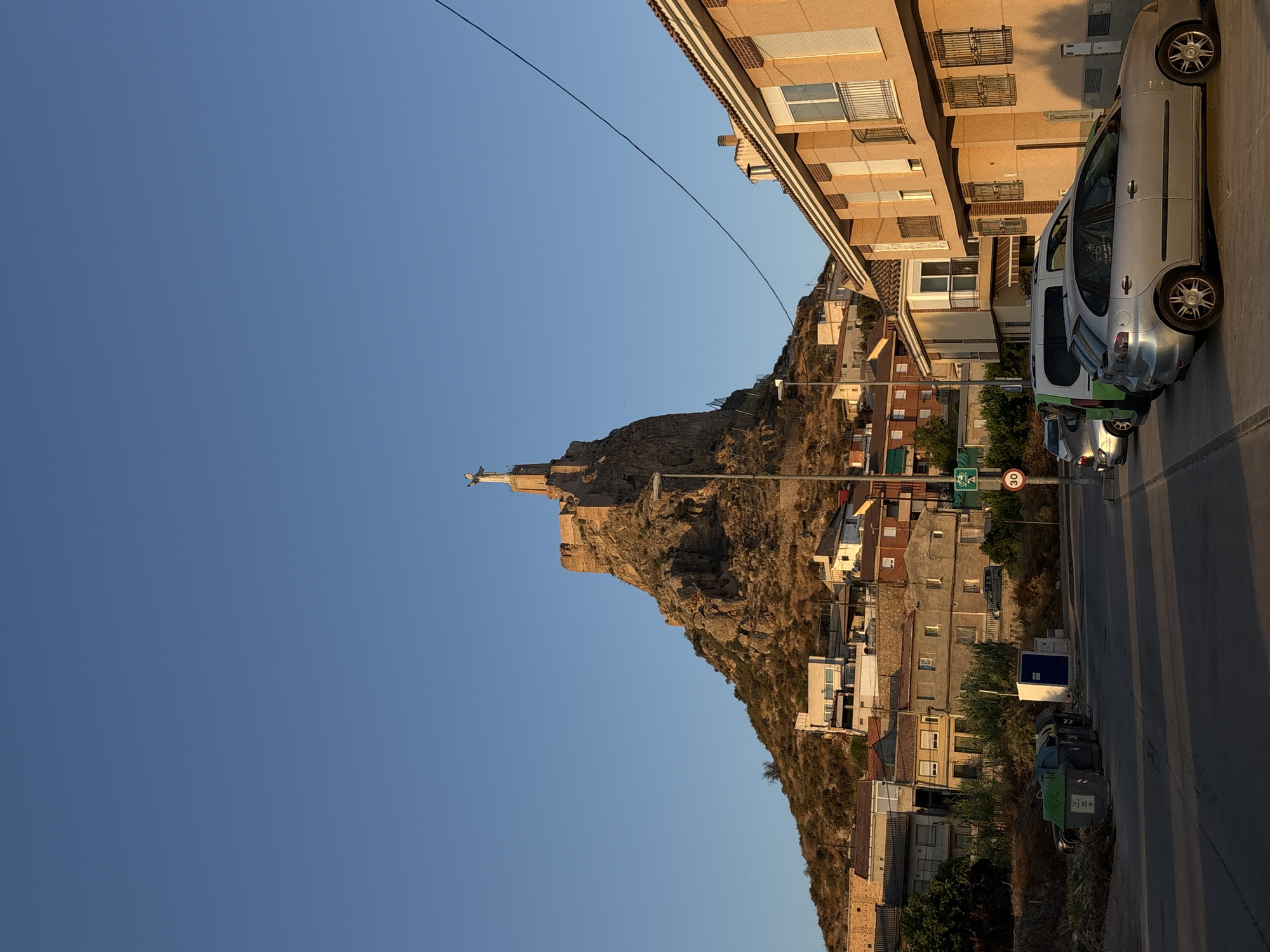 The image shows a view of the Sanctuary of the Virgin of the Castillo in Monteagudo, Spain, from the base of the hill. The sanctuary is perched atop a rocky outcrop, with a prominent statue of Christ the King visible at its summit. The sun is low in the sky, casting long shadows and illuminating the scene with warm, golden light.
In the foreground, a paved street runs horizontally across the frame. Several parked cars are visible on the right side of the street, including a silver hatchback and a white van. To the left of the street, there are rows of municipal waste bins. Beyond the street and bins, the slope of the hill rises, covered with dry vegetation and scattered residential buildings that appear to be built into the hillside.
The buildings in the foreground and mid-ground are typical of a Spanish town, with light-colored facades, tiled roofs, and shuttered windows. A street lamp stands on the right side of the image, near the parked cars. A traffic sign indicating a speed limit of 30 km/h and a bicycle symbol are also visible on the right side of the road.
The sky is a clear, deep blue, indicating fair weather. The overall impression is a peaceful evening scene in a Spanish town, with a significant religious landmark dominating the skyline.