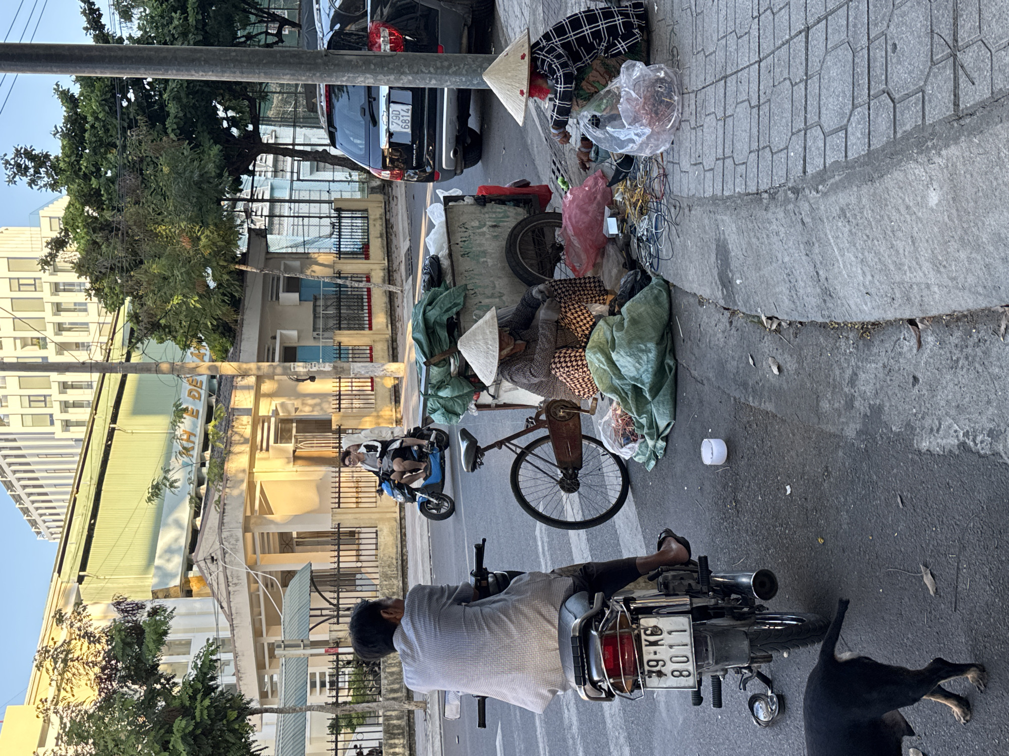 The image depicts a street scene in Nha Trang, Vietnam, likely during daylight given the lighting. In the foreground, a man is seated on a motorcycle, facing away from the camera. The motorcycle has a license plate reading "79-KB 8011". To the right, two individuals, appearing to be women, are seated on the sidewalk. Both are wearing conical hats. The woman on the left is partially obscured by a cart. The woman on the right is engaged in sorting through materials, including wires and what appear to be dried goods in a plastic bag. A bicycle is parked next to the cart. Further back, a blue scooter with two occupants is moving down the street. A black SUV with a license plate "79D 6814" is parked on the right side of the road. Behind the immediate street activity, buildings of varying architectural styles are visible, including a multi-story modern building and a lower building with arched windows and a pitched roof. Trees line the street, and power lines are present overhead. A black dog is visible in the lower left corner of the image. The scene suggests a typical urban environment with street vendors and pedestrian and vehicular traffic.