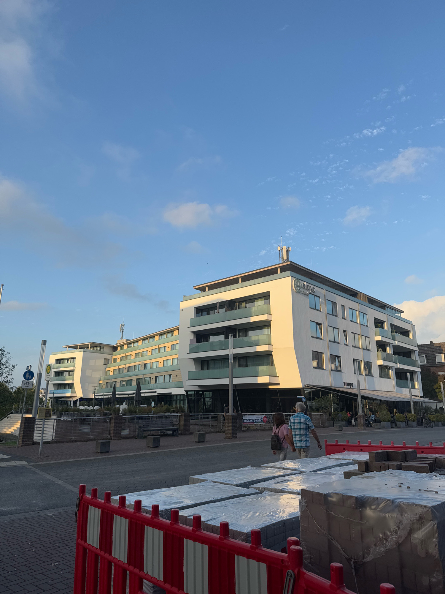 In the city of Cleves, Germany, a modern, white hotel with glass balconies stands under a partly cloudy blue sky. The hotel, identified by the "Dorint Hotel am Niederhein" sign at the top of its facade, features multiple stories and a sleek, contemporary design. In the foreground, a red and white construction barrier is positioned in front of stacks of packaged building materials, possibly paving stones, wrapped in plastic. Two individuals, a man wearing a plaid shirt and shorts, and a woman in a pink shirt and light-colored pants, are walking away from the camera towards the hotel. A street with parked bicycles is visible in the background, along with outdoor seating for a restaurant under a canopy. Traffic signs indicating pedestrian and bicycle paths are also present near the hotel entrance. The scene is illuminated by what appears to be late afternoon sunlight, casting long shadows.