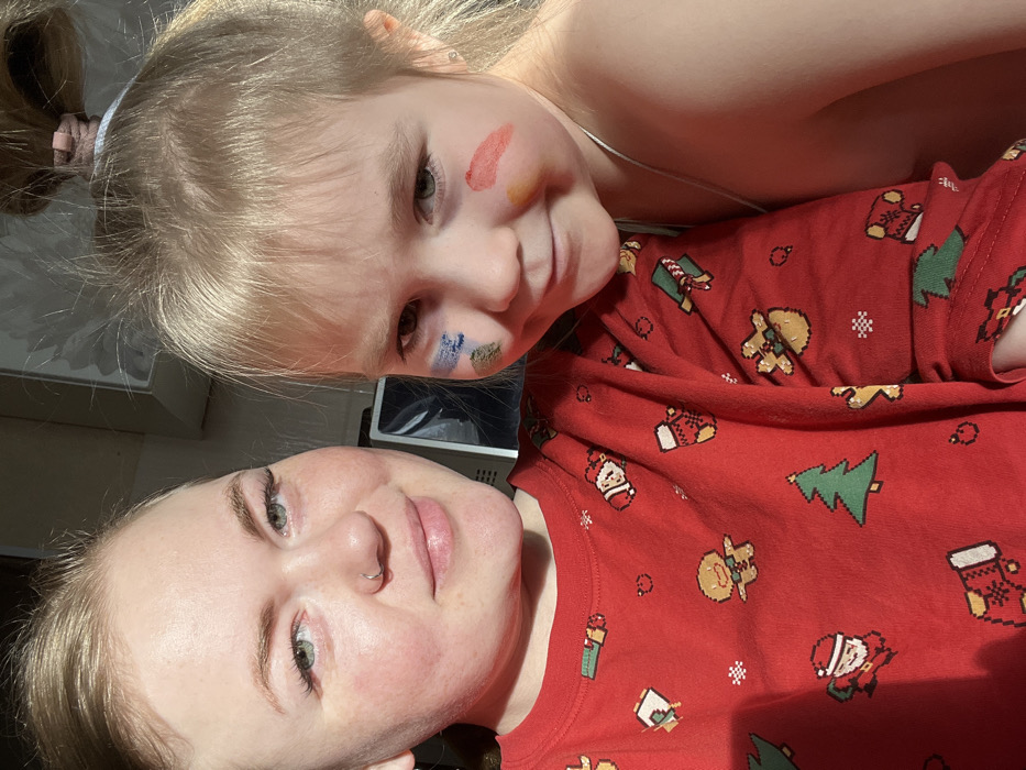 A selfie captures a woman and a young girl close together. The woman is facing the camera directly, wearing a red shirt with Christmas-themed patterns like gingerbread men, trees, and Santa. The young girl is positioned above and slightly behind her, with colorful paint smeared on her face. Both have light skin, and the girl has blonde hair pulled back in a ponytail. The background is blurry and indistinct.