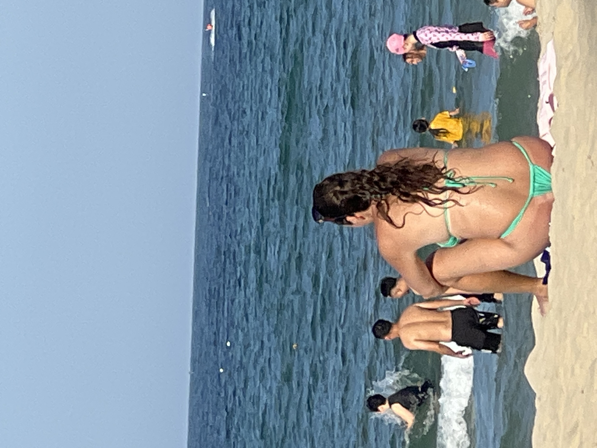 This image depicts a crowded beach scene at Đà Nẵng, Vietnam, with the turquoise waters of the ocean stretching towards a hazy horizon under a clear, pale blue sky. In the foreground, a woman is seated on the sandy shore, facing away from the camera, wearing a green bikini. Her long, dark, curly hair is prominent. To her right, a child wearing a pink cap and a patterned rash guard is partially visible in the water. Further into the sea, several individuals are engaged in various water activities. A person in a yellow shirt is wading, and to the left, two men are in the shallow water, with one splashing. In the distance, a white jet ski is being ridden by a person. The overall atmosphere is one of a typical, busy summer day at the beach.