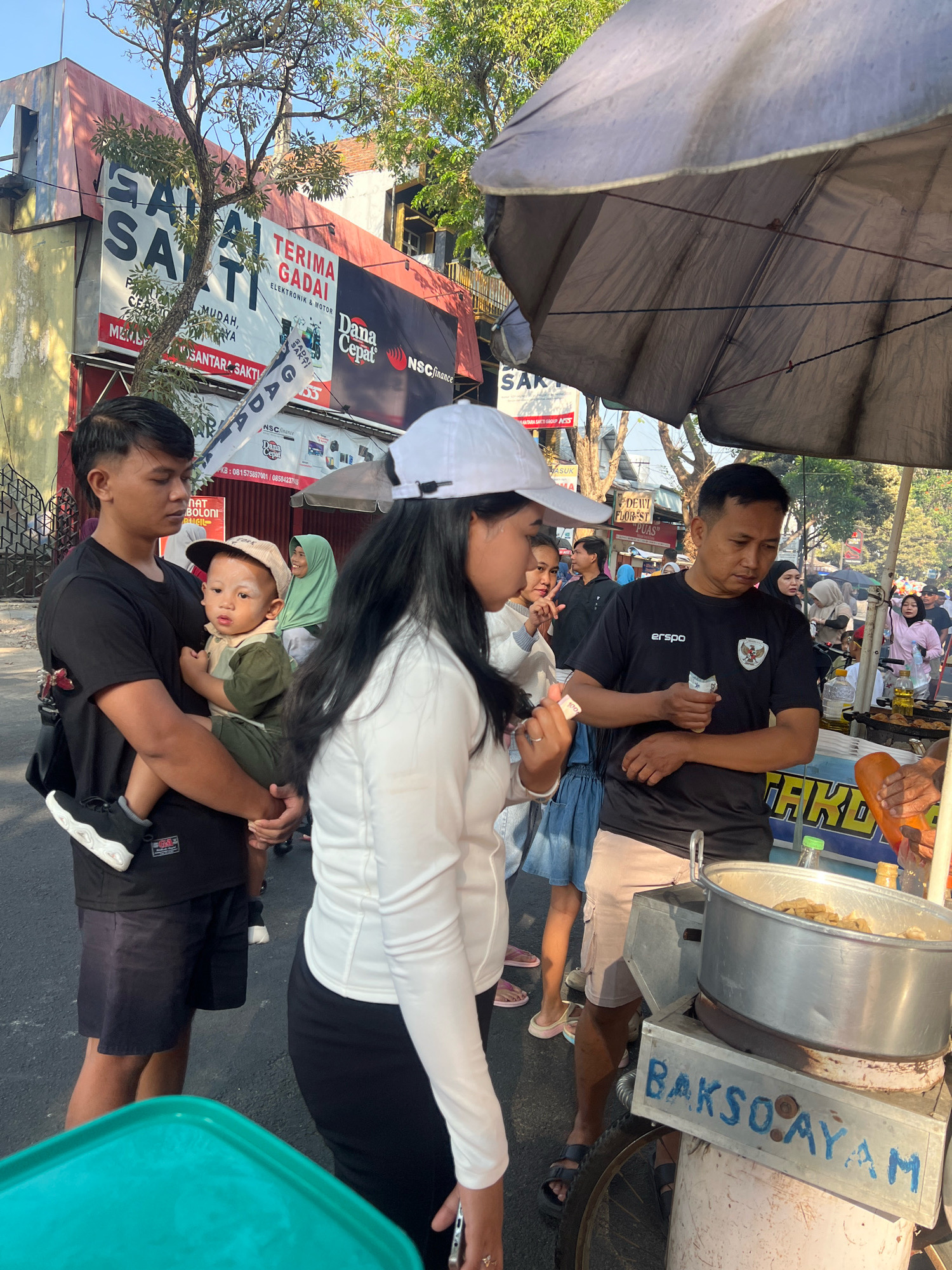 A man in a black t-shirt holds a toddler in his arms. The toddler wears a green shirt and a beige cap. A woman in a white long-sleeved shirt and a white cap stands to the right of the man, facing away from the camera. Behind them, a food cart with a large pot of food is visible, labeled "BAKSO AYAM". A man in a black t-shirt with a logo is attending the food cart and appears to be interacting with the woman. In the background, there are buildings with various signs, suggesting a street-side commercial area. The scene appears to be outdoors, with sunlight casting shadows. The location is identified as Kecamatan Pati, Indonesia.