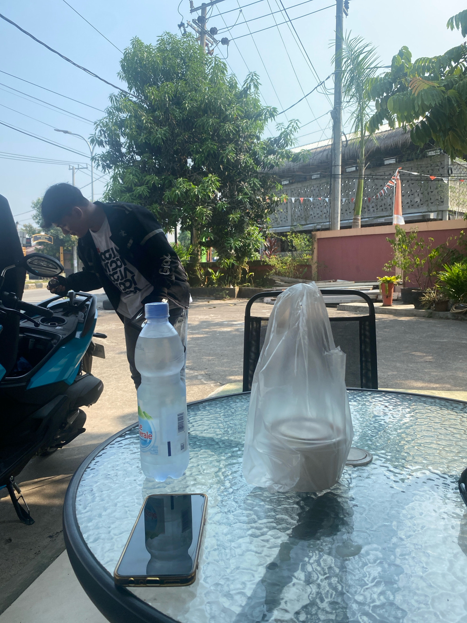 A young person wearing a black jacket and a white t-shirt is seen leaning over a teal motorcycle parked on a paved area. In the foreground, a glass-top table holds a bottle of water and a mobile phone. Another object, possibly food in a takeaway container, is covered with a plastic bag on the table. The background features trees, buildings with thatched roofs, and utility poles, suggesting an outdoor setting in a semi-urban or suburban area, likely in Tanjung Redeb, Indonesia, given the cultural context suggested by the Indonesian flag banner and architectural style. The scene appears to be captured during daylight hours under a clear sky.