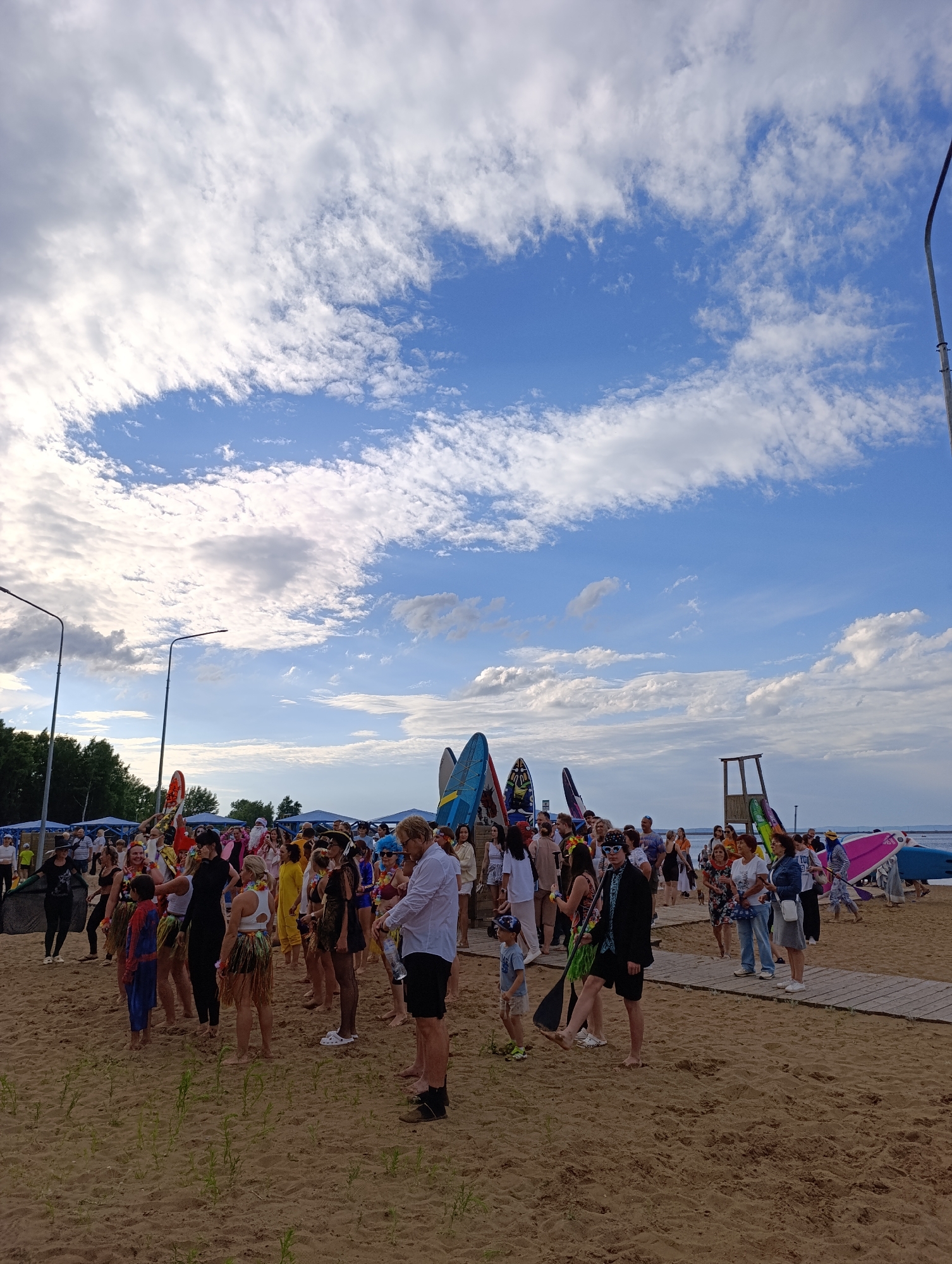 A group of people are gathered on a sandy beach, with a body of water visible in the background. Many of the individuals are dressed in colorful, festive attire, including leis, Hawaiian skirts, and some costumes. Several surfboards and paddleboards are standing upright, some adorned with decorative patterns. Tents are set up further back on the beach, and trees line the horizon. Streetlights are also present. The sky is a bright blue with scattered white clouds, suggesting it is daytime, likely late afternoon or early evening given the soft lighting. The overall atmosphere appears to be a celebratory event or gathering, possibly related to water sports or a summer festival. Some people are walking, others are standing and talking, creating a lively scene. There is a wooden lifeguard tower visible to the right of the frame. The ground is sandy with some sparse green vegetation growing.