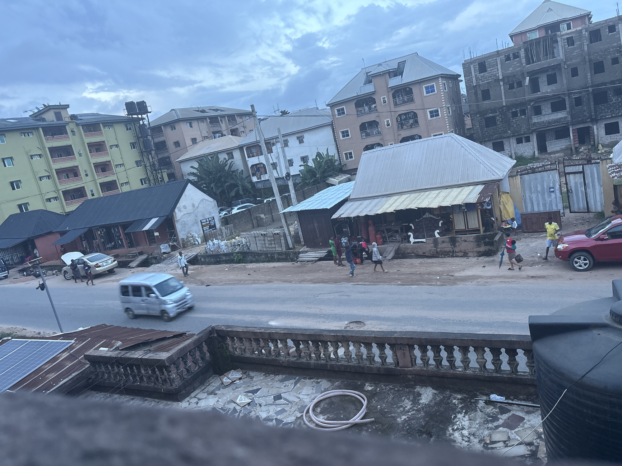 The image depicts a street scene in Nkpor, Nigeria, under a cloudy sky. A light-colored minivan is in motion on the asphalt road, with its headlights on. To the left of the road, there is a building with a green facade and several floors, featuring balconies and numerous windows. Adjacent to it, a dark-roofed structure is visible. Further down the road, a red SUV is parked.
Several individuals are present on the sidewalk and road. One person in a yellow shirt stands near the red SUV. Further left, a group of people are gathered near a building with a light-colored corrugated metal roof, where some are interacting with what appear to be products or wares. Two people are walking on the sidewalk, and another is seen walking with an umbrella.
In the foreground, a low, decorative concrete wall with ornate railings separates the street from an elevated area. This area is paved with broken tiles and has a coiled white hose lying on the ground. A portion of a large black water tank is visible on the far right of the foreground. A solar panel is mounted on a corrugated metal roof to the left. The background features multiple buildings of varying heights and architectural styles, some appearing to be under construction with exposed brickwork.