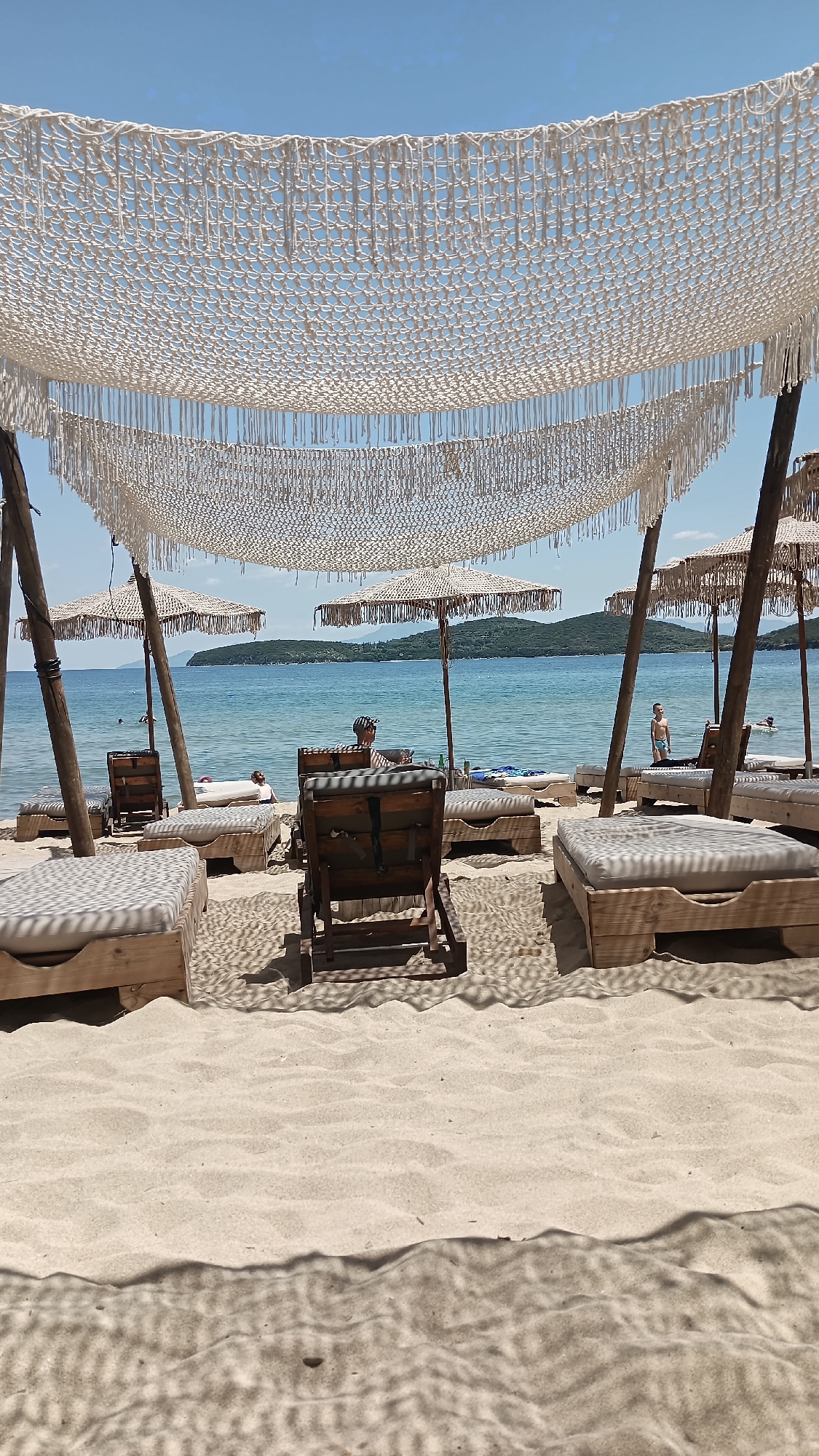 The image captures a serene beach scene on a sunny day, likely in Nea Peramos, Greece. The foreground is dominated by a sandy beach with light, rippled sand casting intricate shadows from the overhead canopies. Several wooden lounge chairs with comfortable cushions are arranged on the sand, some occupied by people.
In the mid-ground, the calm, blue sea stretches out towards a lush, green, tree-covered island in the distance. The water is clear, and a few people can be seen enjoying the ocean, with one person swimming and another standing near the shore. Several beach umbrellas, made of straw or similar natural material, provide shade, and a distinctive macrame-like canopy stretches across the top of the frame, adding a bohemian touch to the setting.
Two individuals are prominently featured relaxing on lounge chairs. One person, wearing a cap, is seated and appears to be looking towards the sea. Another person, a child, stands near the water's edge, looking out at the ocean. The atmosphere is peaceful and relaxed, suggesting a vacation or leisure activity. The bright sunlight indicates it is likely midday. No visible text is present in the image.