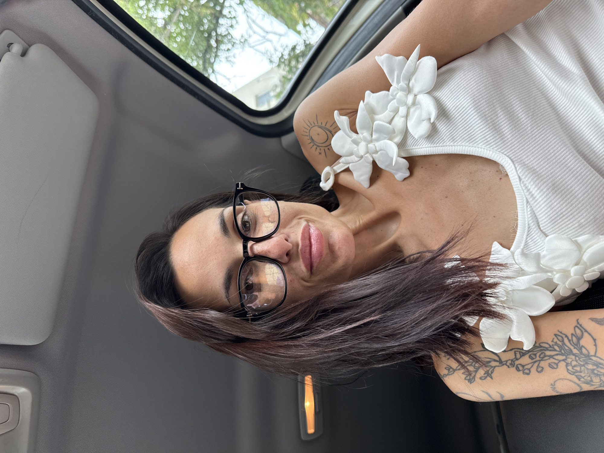 A woman with dark brown hair and glasses is taking a selfie in a car. She is wearing a white tank top with floral appliques on the straps. She has tattoos on her left arm and shoulder. The car interior is visible, with the sun visor and part of the ceiling. Outside the car window, trees and a building are visible. The image was taken in Pleven, Bulgaria.