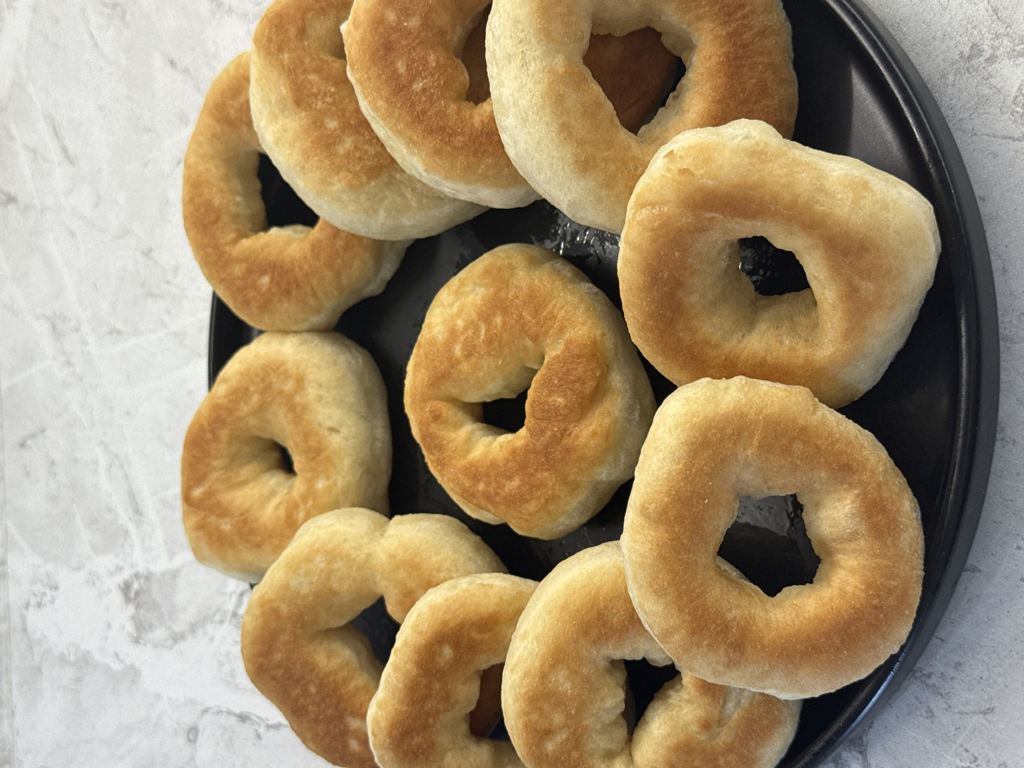 A black plate filled with freshly baked, golden-brown bagels sits on a textured gray and white surface. The bagels are arranged in a circular pattern, with some overlapping. They appear to be small, bite-sized, and some have a slightly irregular shape, suggesting they are homemade. The lighting is bright, casting soft shadows and highlighting the texture of the bagels. There is no visible indication of the location being in Lyubertsy, Russia; the background appears to be a kitchen or dining area.
