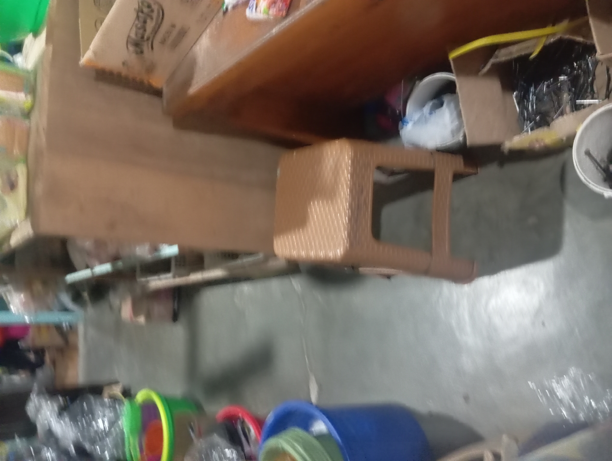 The image shows a low-angle, slightly out-of-focus view of what appears to be a storage area or a small shop in Rejosari, Indonesia. The floor is concrete, with some scuff marks and what looks like a wet patch on the left side.
In the foreground on the left, there are several plastic buckets. The largest is a bright blue bucket, containing green and red smaller buckets or containers. To the left of the blue bucket, there is a red bucket with its contents unclear. Further left, out of focus, are more items, some appearing to be wrapped in plastic.
Dominating the right side of the image is a wooden structure, possibly a counter or shelf unit, with a light brown finish. On top of this structure, there are several items including a box with the word "Masako" visible, a brand of seasoning in Indonesia, and other indistinct packages.
In the middle ground, to the right of the buckets and in front of the wooden structure, is a small, brown plastic stool with a woven pattern. Behind the stool, there are several cardboard boxes. One box contains a white plastic bag, and another is filled with dark, metallic-looking objects, possibly nails or screws, with yellow plastic straps visible.
In the background, the scene is very blurry, but it suggests a cluttered environment with more shelves and items. The overall impression is of a functional, perhaps slightly disorganized, commercial or storage space. No people are clearly visible.