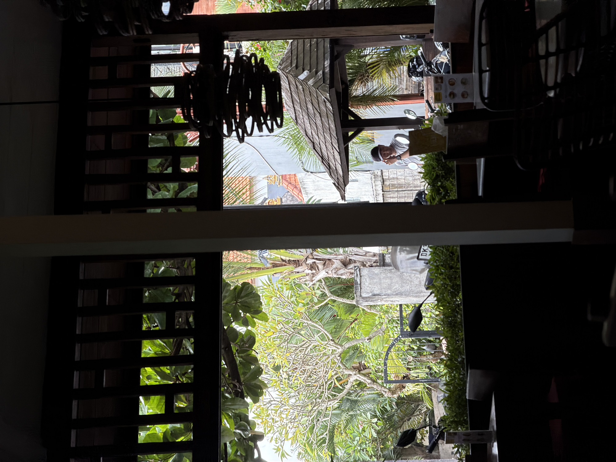 The image, rotated ninety degrees, shows a view from inside a building, likely a cafe or restaurant, looking outwards towards a street in North Kuta, Indonesia. A slatted wooden structure and a hanging chandelier frame the view.
Outside, a person in a white shirt and dark cap is standing near a parked motorcycle. Further down the street, the roofs of buildings are visible amidst lush green foliage, including palm trees.
A concrete wall and a sign, possibly a menu board, are also visible. The weather appears to be bright and sunny.