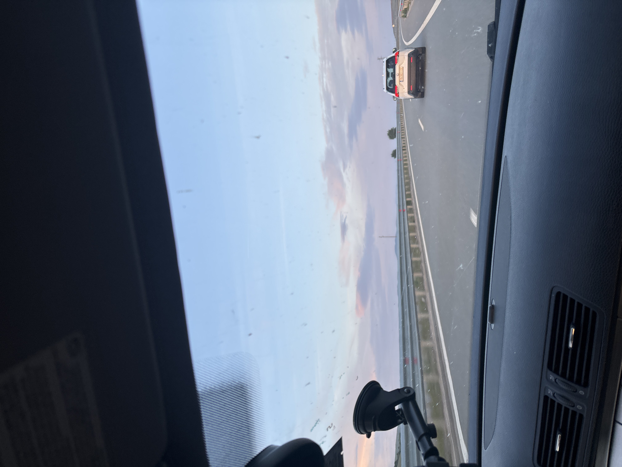 The image shows a view from inside a car driving on a highway in Aldomirovtsi, Bulgaria. A white car is visible in the distance ahead, traveling in the same direction. The sky is visible through the windshield, displaying a mix of blue and sunset colors. A suction cup mount is affixed to the windshield, holding a device that is partially visible. The car's dashboard and air vents are visible in the foreground. Roadside barriers are seen along the highway.