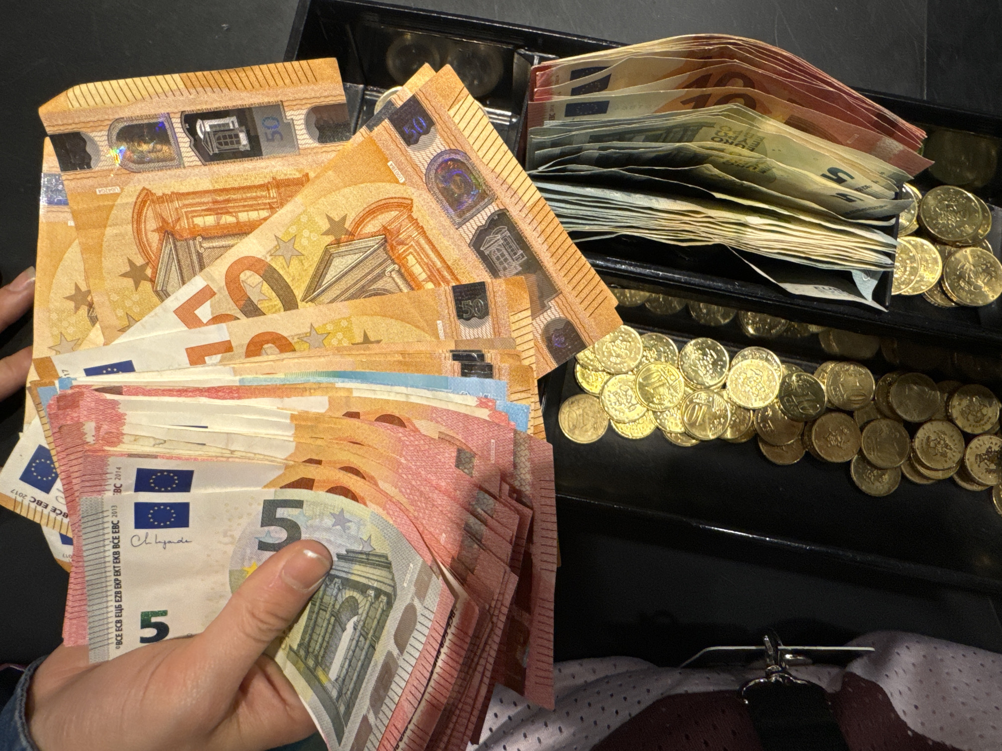 A close-up shows a cash register drawer filled with Euro currency in an unknown city. The drawer contains stacks of banknotes, including 5 Euro and 50 Euro bills. A hand holds a collection of 5 and 50 Euro notes, further demonstrating a monetary transaction. Additionally, a collection of 10 cent Euro coins is present in the drawer.