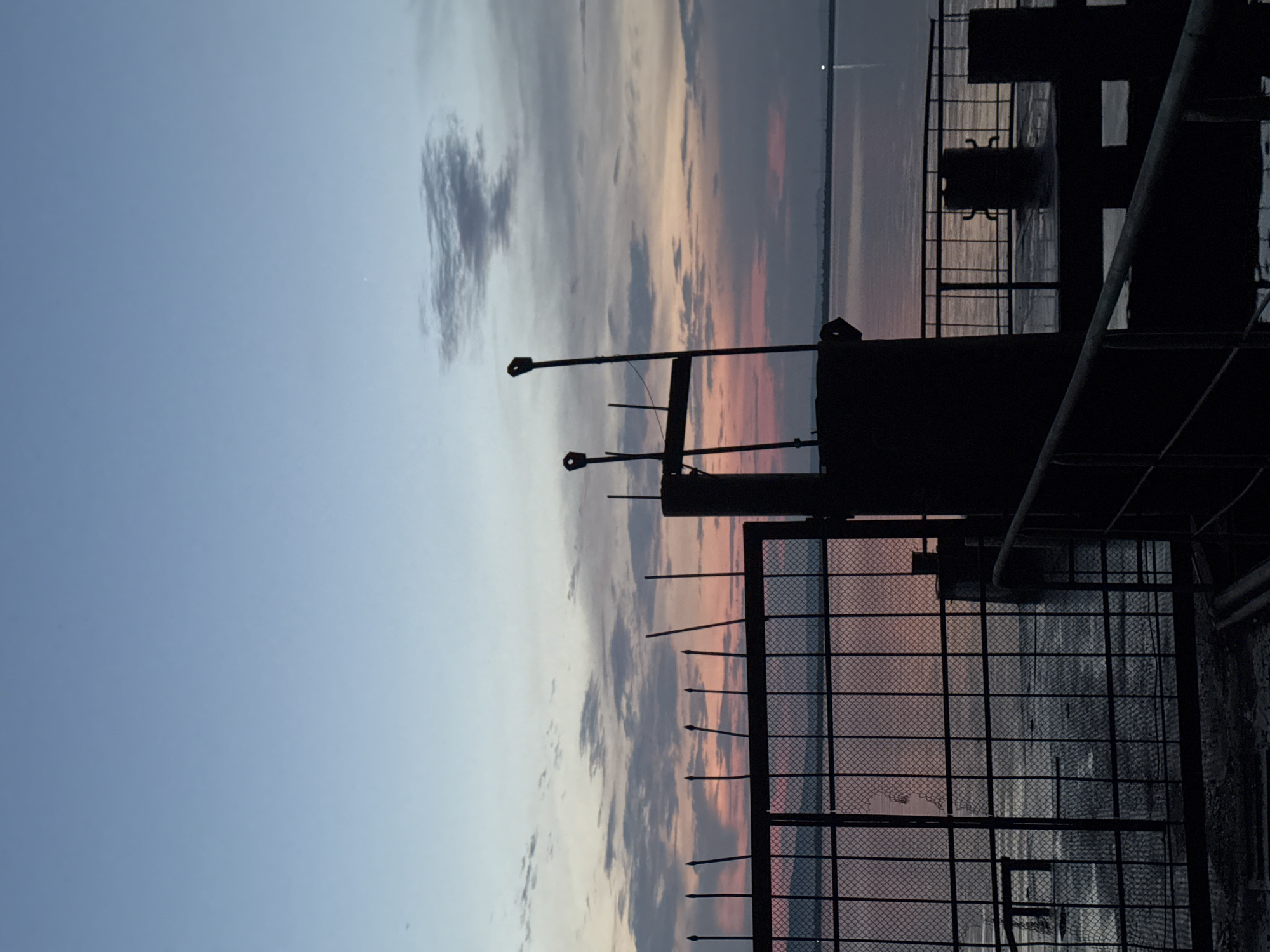 This is a silhouette photograph taken at dusk, possibly from the embankment in Chaikovsky, Russia. In the foreground, a dark metal fence with pointed spikes runs horizontally across the frame. Behind the fence, a dark, industrial-looking structure with two vertical poles with rings at the top is visible. To the right of this structure, a section of a railing and a bollard are also silhouetted. The background features a body of water, likely the Kama River, reflecting the colors of the sunset. The sky above is a gradient of pale blue at the top, transitioning to hues of pink, orange, and gray clouds as it approaches the horizon. Distant, indistinct landmasses or structures are visible along the far edge of the water. The overall impression is one of industrial waterfront infrastructure against a dramatic natural backdrop.