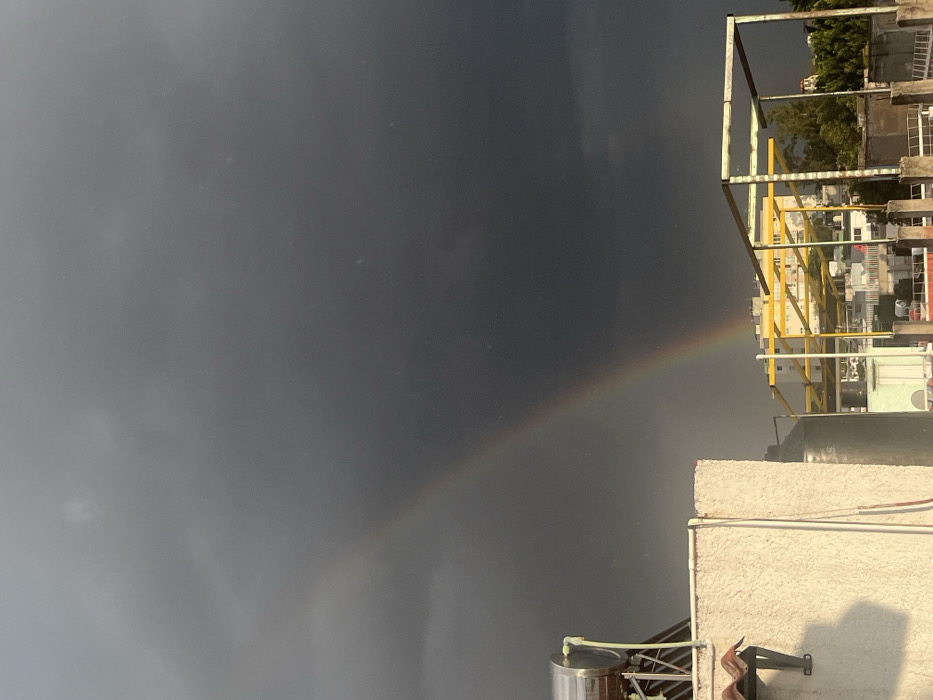 The image displays an elevated view of an urban landscape in the city of Unknown, Unknown, under a dark gray, heavily overcast sky. A prominent rainbow arcs across the upper half of the frame, originating from the lower left sky and extending towards the mid-right, passing over distant structures. In the lower left quadrant, a section of a white-painted building or rooftop is visible, featuring a silver cylindrical water tank or solar heater mounted on a metal support frame. Below this, a portion of a reddish-brown tiled roof overhang is present. In the mid-ground, a large, rectangular, yellow-painted metal framework extends horizontally across multiple lower rooftops. Various residential or commercial buildings of differing heights and green foliage are visible between and beyond the foreground structures. No individuals are present in the scene.