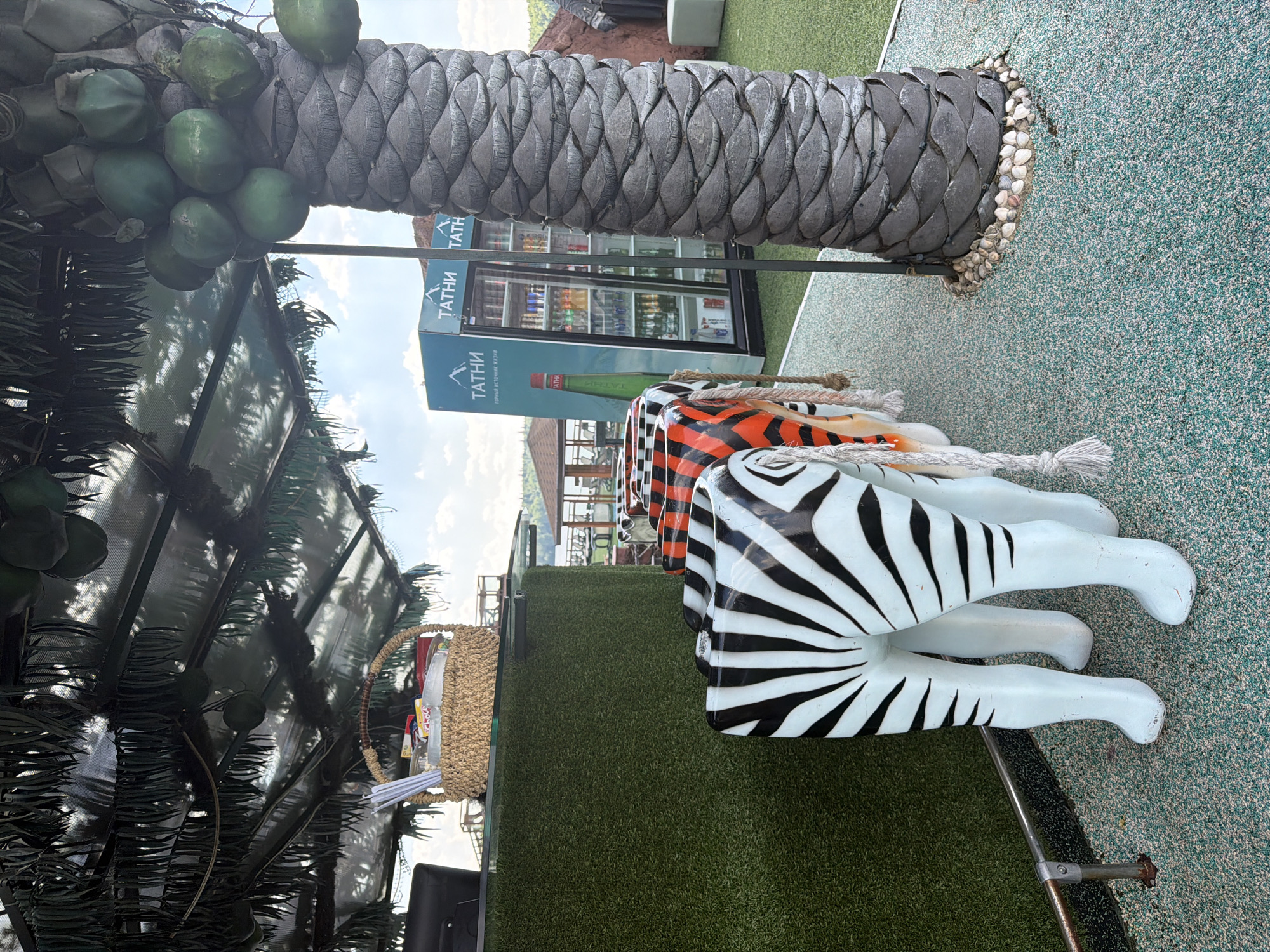 A row of zebra-striped chairs, shaped like the hindquarters of zebras with their legs forming the chair legs, are lined up next to a counter covered in artificial grass. A basket filled with straws and packets sits on the counter. To the right of the chairs, a large artificial palm tree stands prominently. In the background, a refrigerator stocked with drinks is visible, along with a large green bottle of water. The scene appears to be an outdoor seating area, possibly at a restaurant or bar, with a backdrop of a cloudy sky.