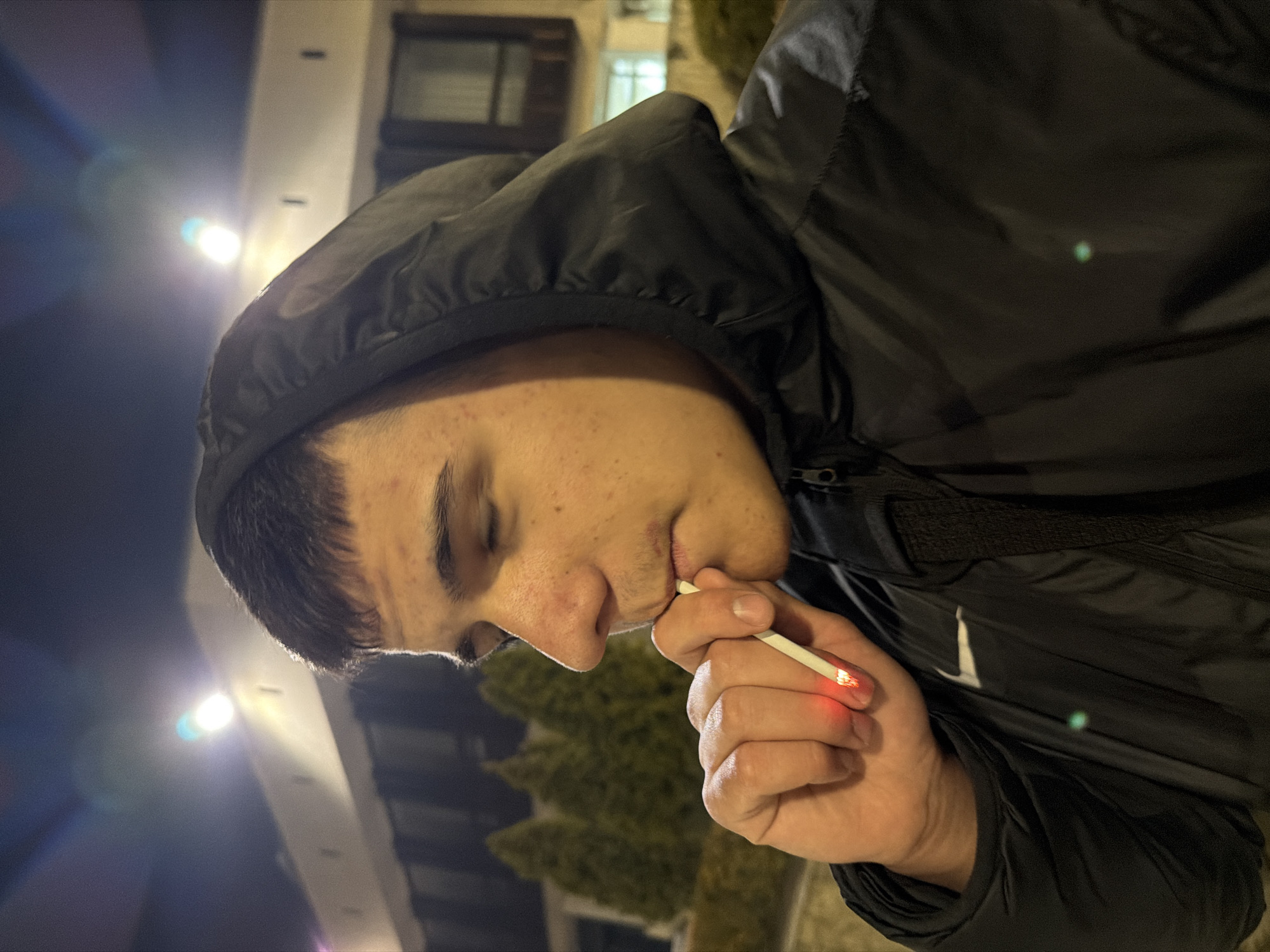A young man with short dark hair and acne on his face is smoking a cigarette. He is wearing a black hooded jacket. The background shows a building lit by streetlights and some greenery. The image was taken at night in what is possibly the city of Panagyurishte, Bulgaria.