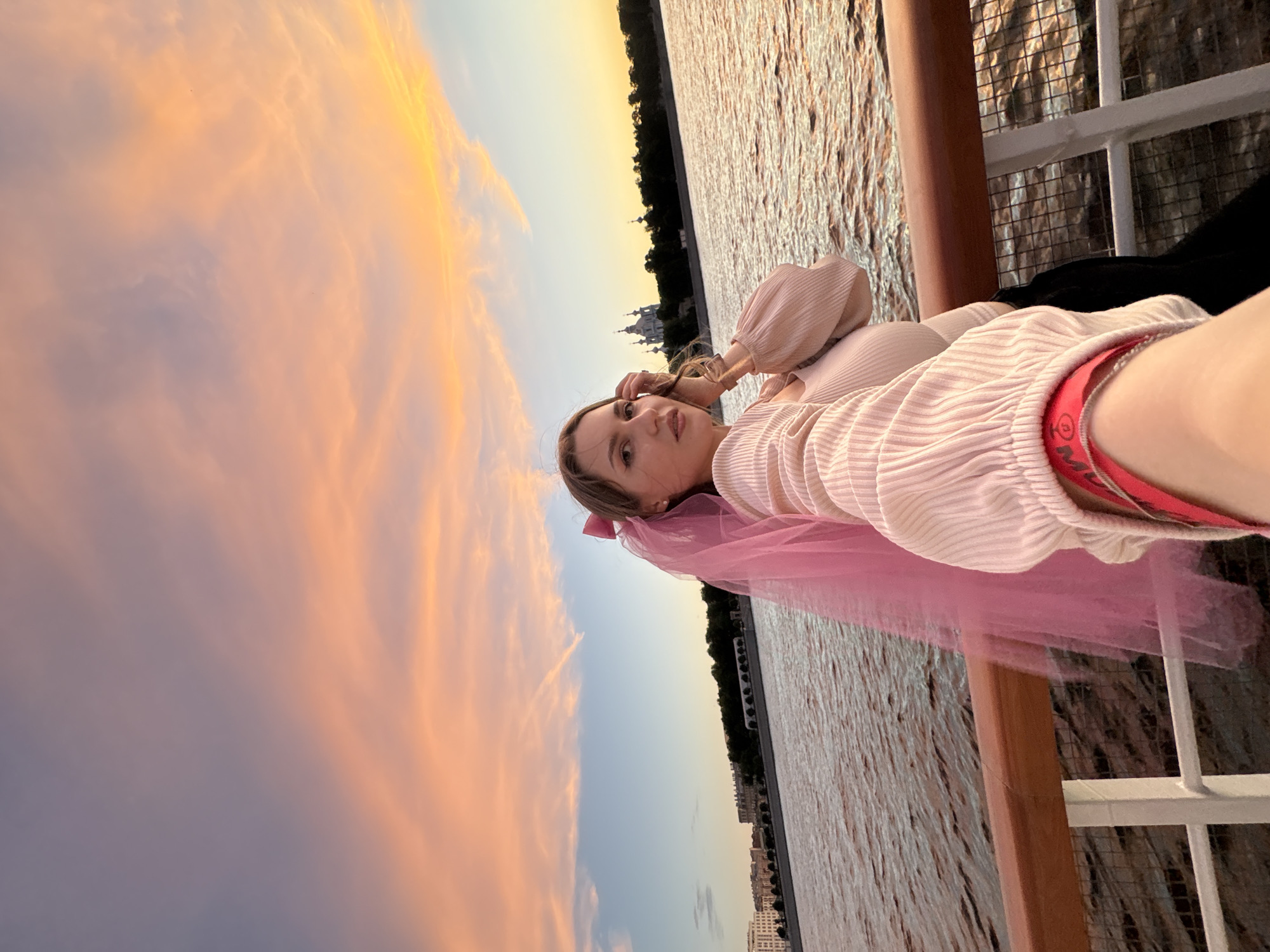 A woman with a pink veil and bow is taking a selfie on a boat at sunset. She is wearing a light-colored, ribbed blouse with puffed sleeves. The boat has a wooden railing and white metal posts. The background shows a body of water, a cityscape with buildings, and a church with multiple domes. The sky is filled with vibrant pink and orange clouds. The setting appears to be the Neva River in Saint Petersburg, Russia.