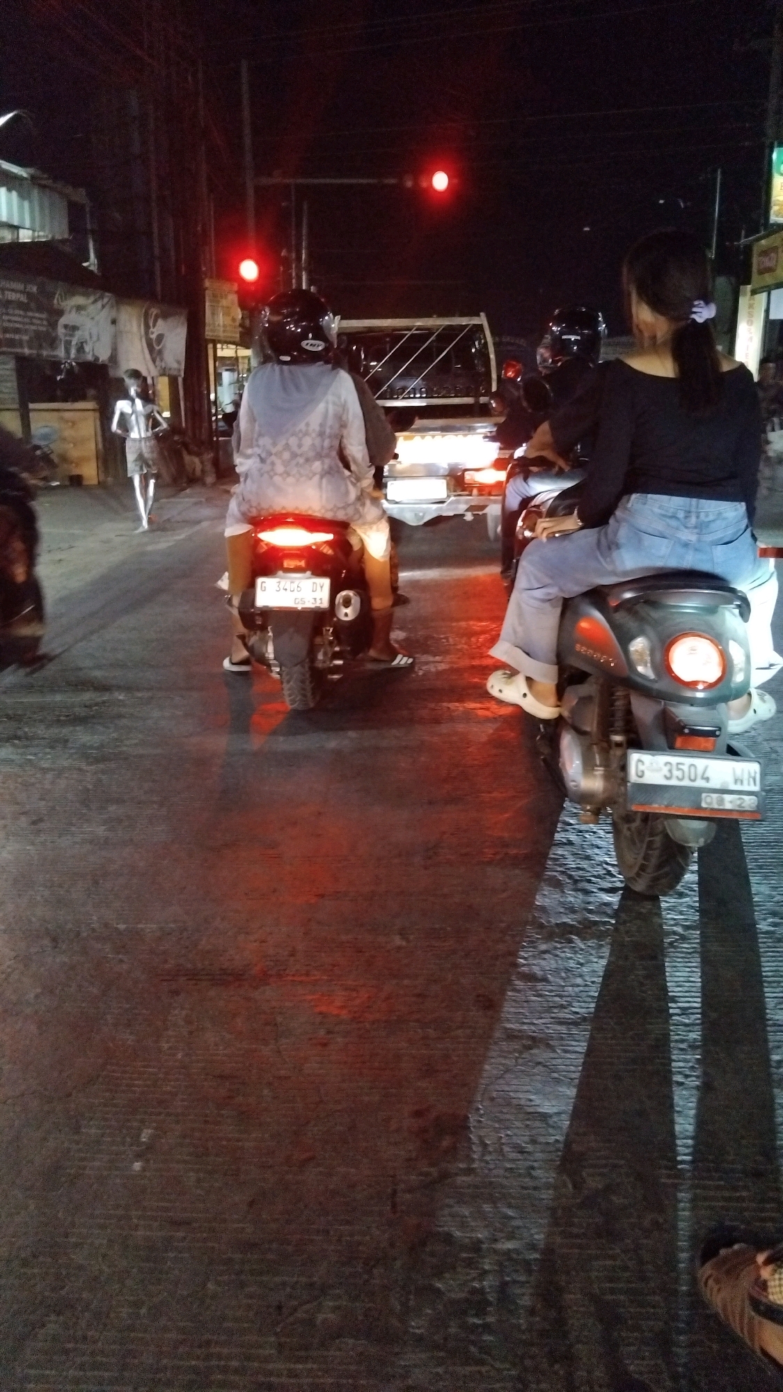 The image shows a night scene in Debong Wetan, Indonesia, at a traffic intersection. Two motorcycles are stopped at a red traffic light. On the left motorcycle, a person wearing a white hoodie and light-colored pants is seated as the rider. On the right motorcycle, a person wearing a black off-the-shoulder top and blue jeans is seated as the passenger, with their arms around the rider. The rider of the right motorcycle is wearing a black helmet. The road surface is wet and reflects the red light from the traffic signal, creating a reddish-orange hue. In the background, a white truck is also stopped at the intersection. To the far left, a person in a light-colored shirt and shorts is standing on the sidewalk. Buildings and streetlights are visible in the background, indicating an urban environment.