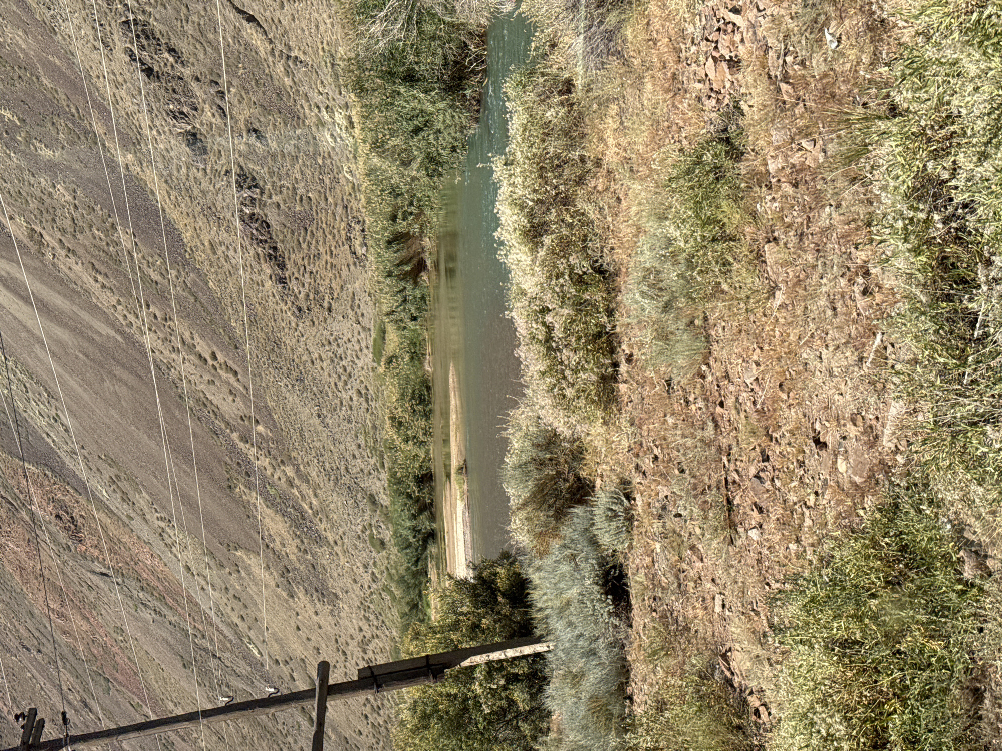 The image depicts a natural landscape dominated by a river flowing through a valley. The river is flanked by lush green vegetation on both sides. In the foreground, dry, rocky terrain is covered with sparse, yellowish-brown grass and low-lying shrubs. A wooden utility pole stands to the left of the frame, with several wires extending from it across the scene. The background features a steep, barren mountainside with striations of various earthy colors, predominantly brown and reddish-brown, indicating geological layers. The sky is not visible in the frame. The scene appears to be in a rural or remote area, possibly a national park or a sparsely populated region, given the absence of any visible structures or human activity beyond the utility pole. The time of day is clearly daytime, with strong sunlight casting shadows.